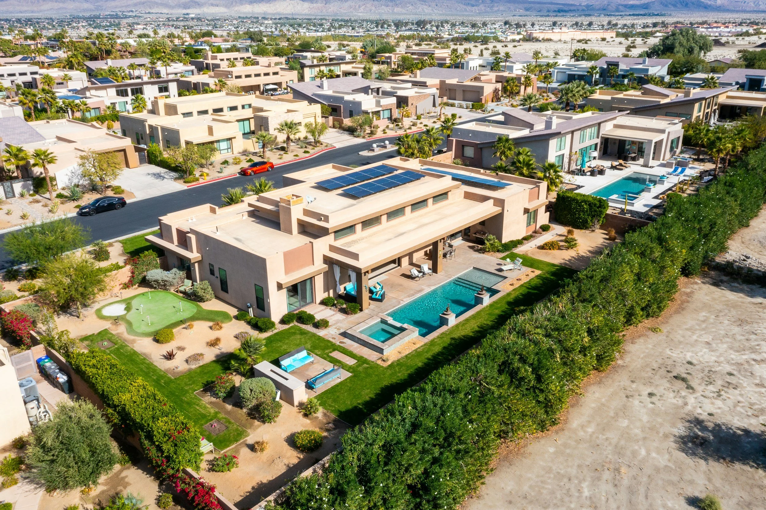 Luchtfoto van een modern huis met een tuin, zwembad en zonnepanelen op het dak, omgeven door een wijk met vergelijkbare huizen en een groene haag.