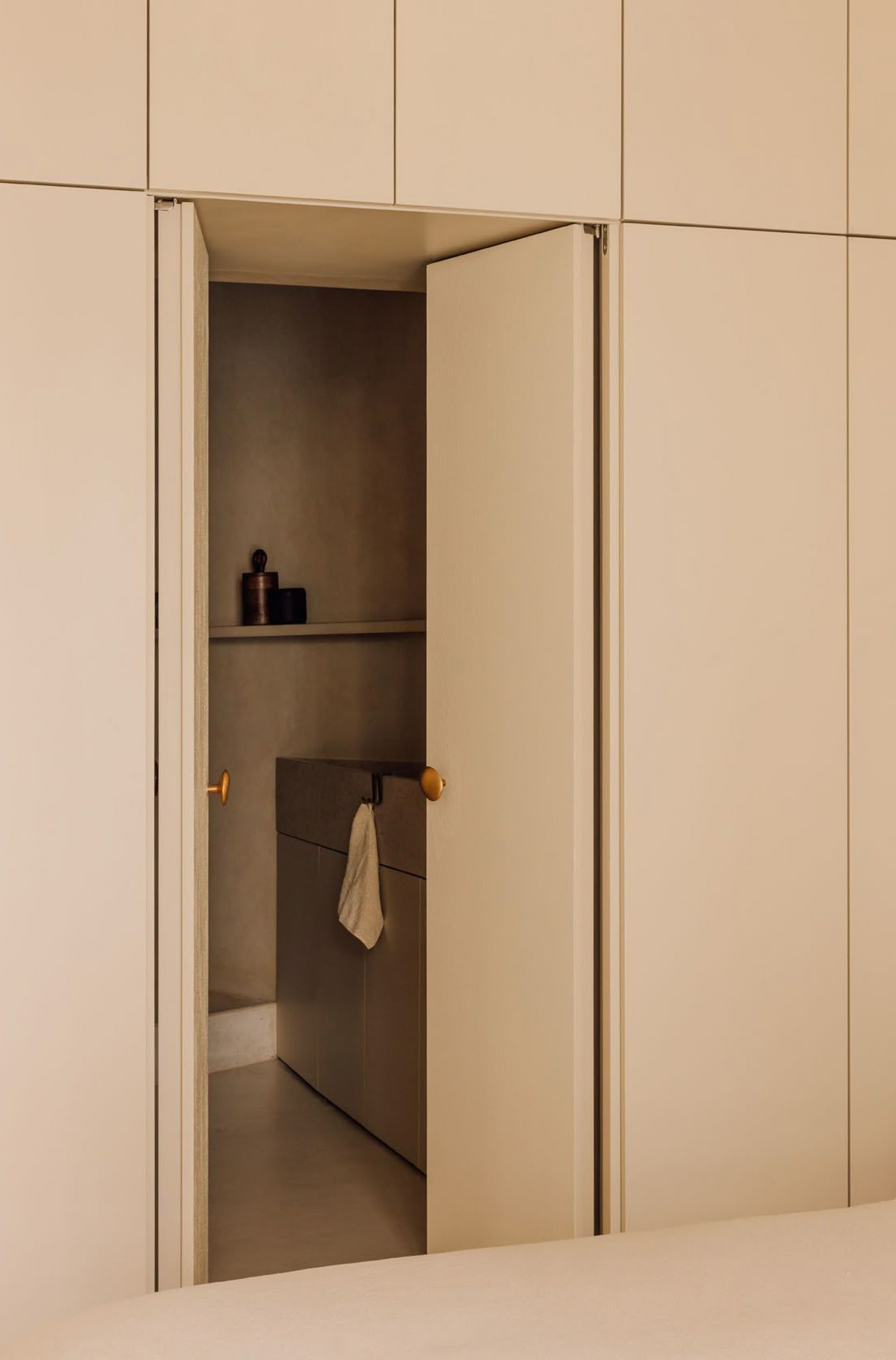 Een open kastdeur die toegang geeft tot een kleine ruimte met een wastafel, een handdoek hangt eraan, en een paar decoratieve voorwerpen op een plank.