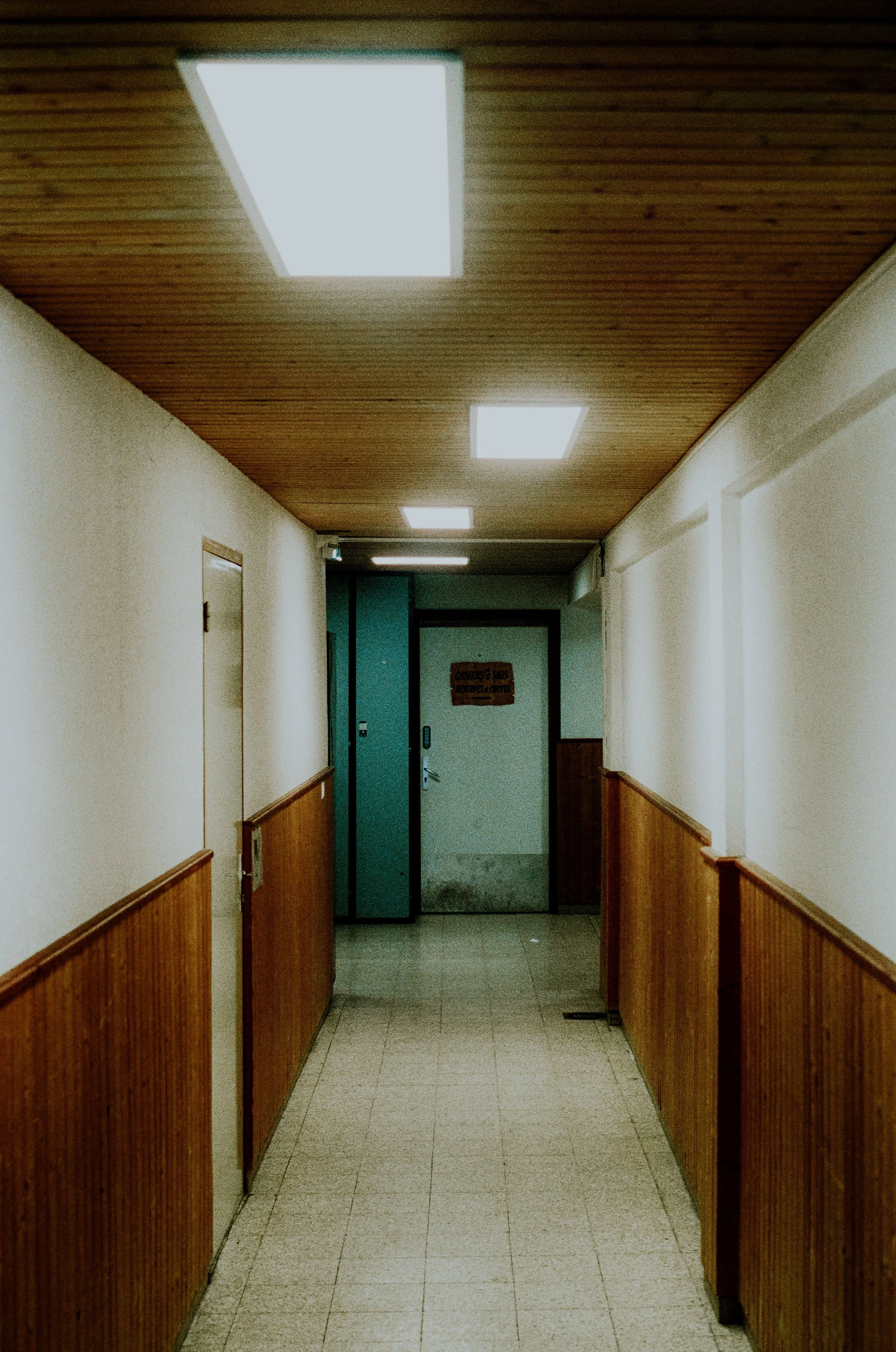 Empty hallway with wooden paneling on the walls, ceiling, and fluorescent ceiling lights, leading to a door at the end.