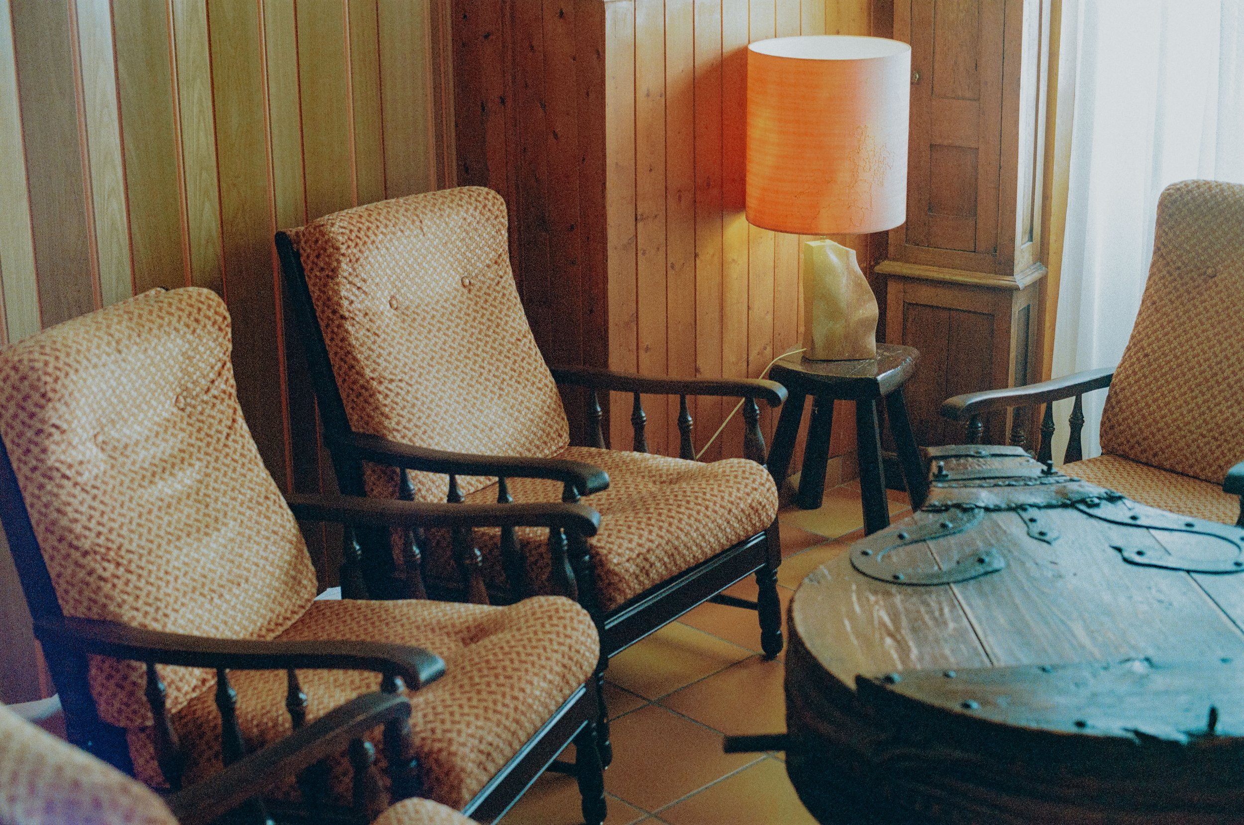 A cozy living room corner with wooden panel walls, featuring three vintage armchairs with patterned cushions, a wooden coffee table with metal accents, and a table lamp with an orange shade on a small side table.