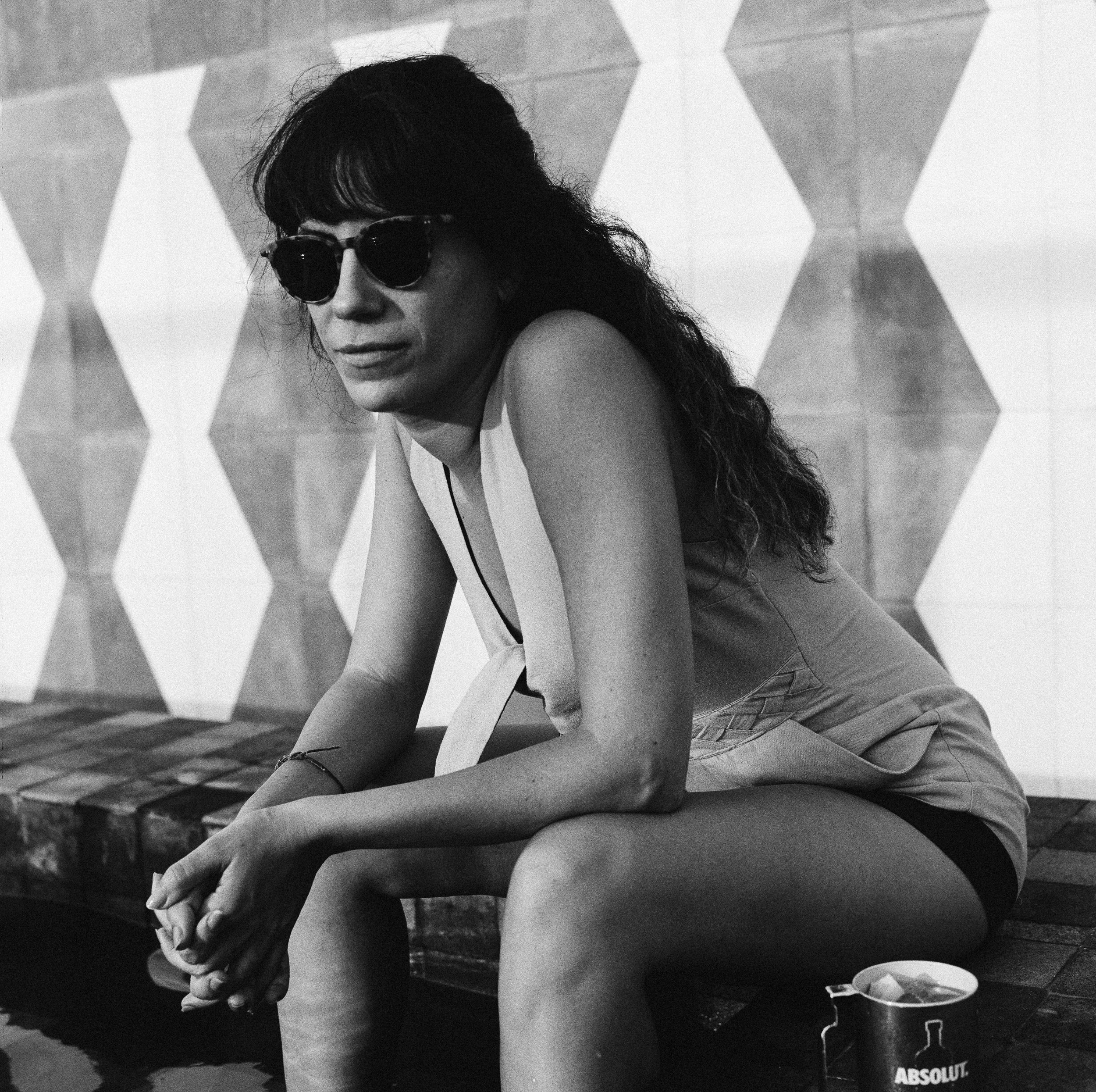 A woman wearing sunglasses, a sleeveless top, and shorts sitting on a wooden bench in front of a hexagonal patterned wall, with a can of Absolut vodka nearby.