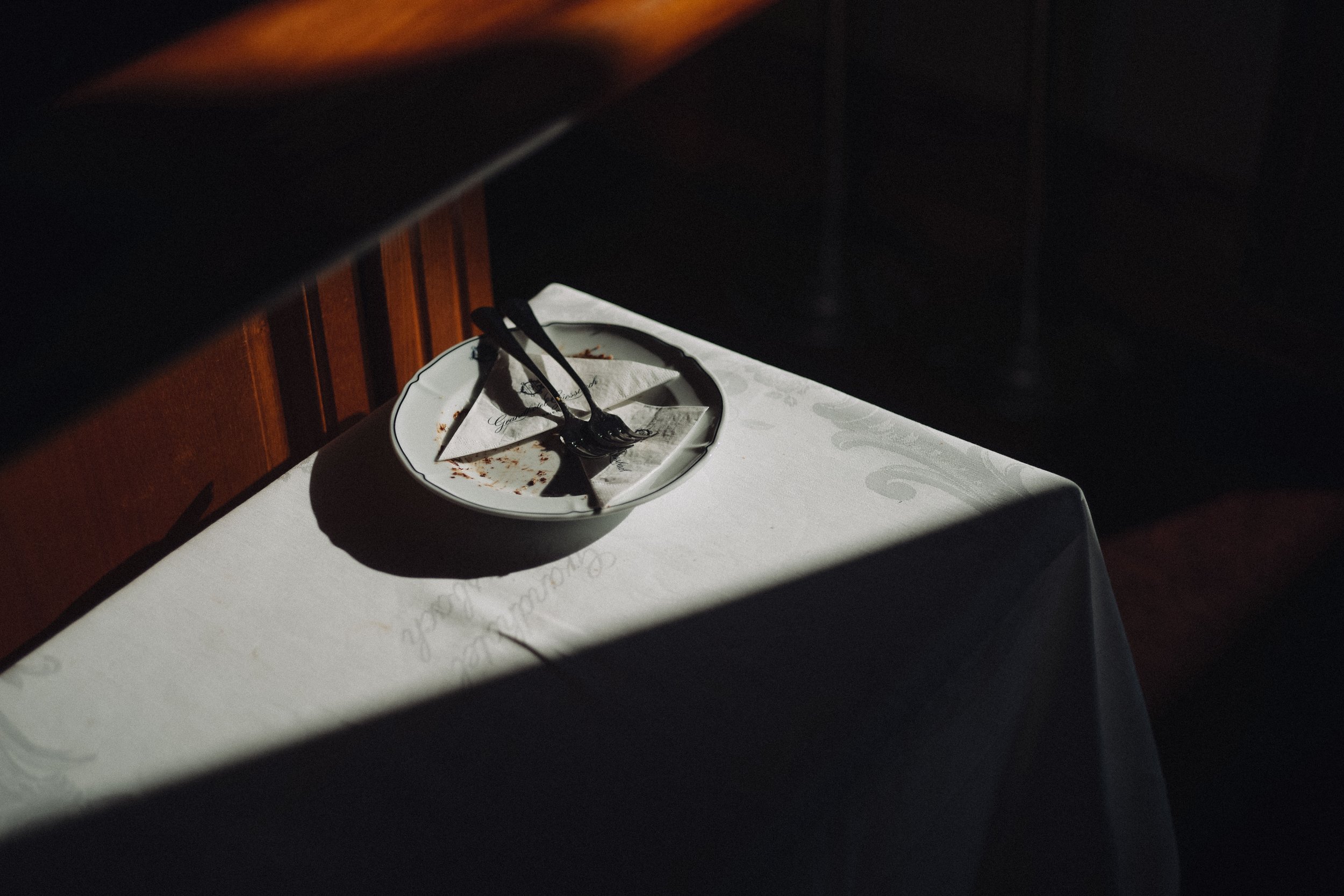Empty white tablecloth-covered table with a white plate, a folded napkin, and three utensils in a dimly lit restaurant.