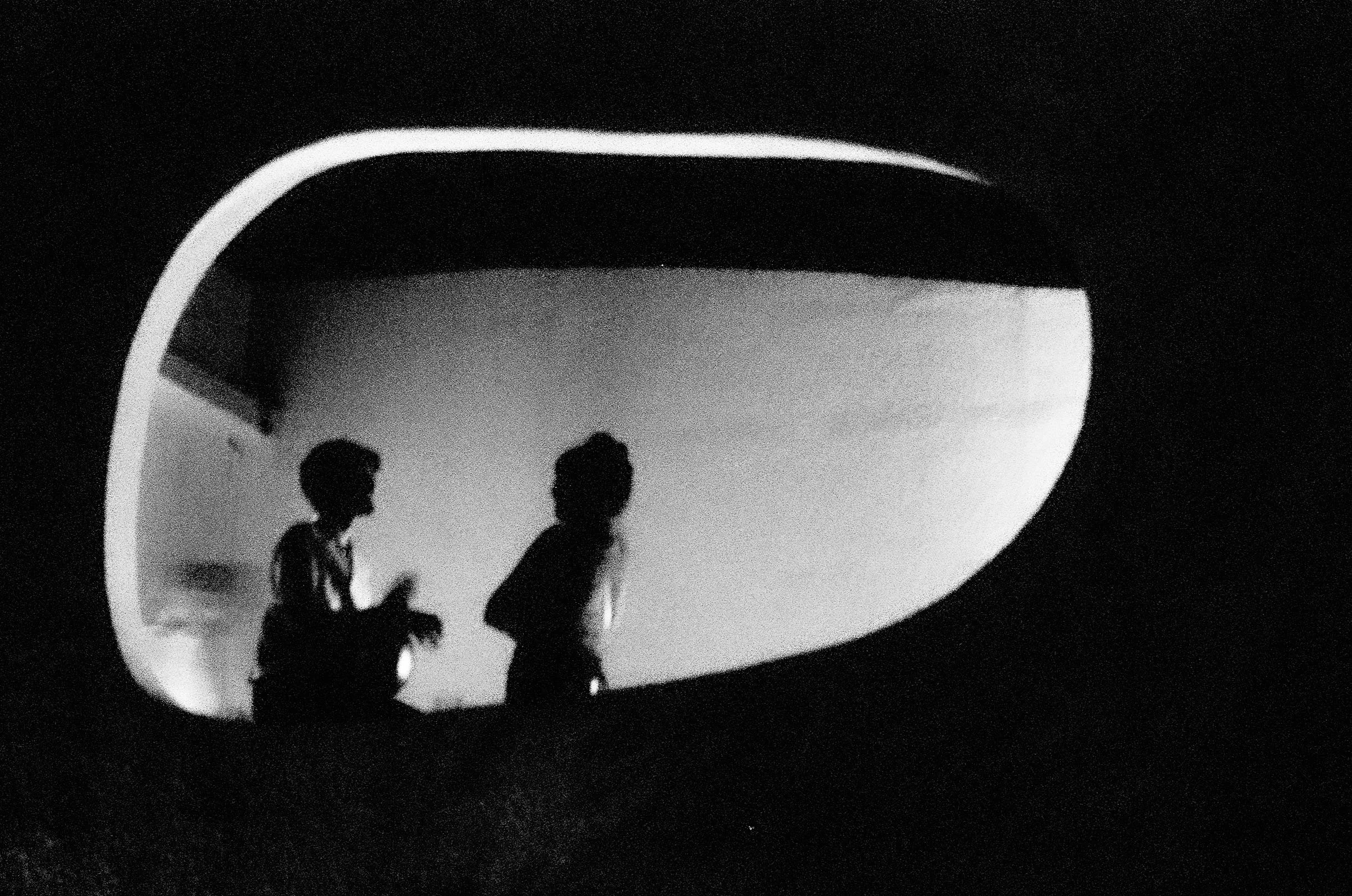 Black and white photo of two children with backpacks, seen through an oval-shaped window with a bright edge, likely in a structure or vehicle.
