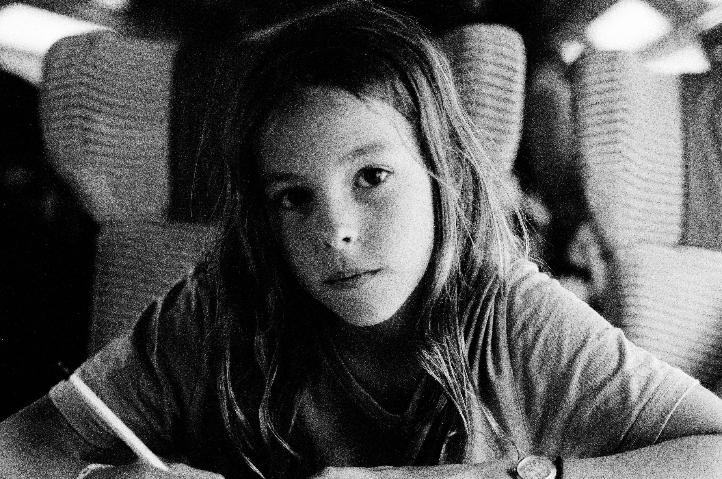 Black and white photo of a young girl with wavy hair looking at the camera, sitting at a table.