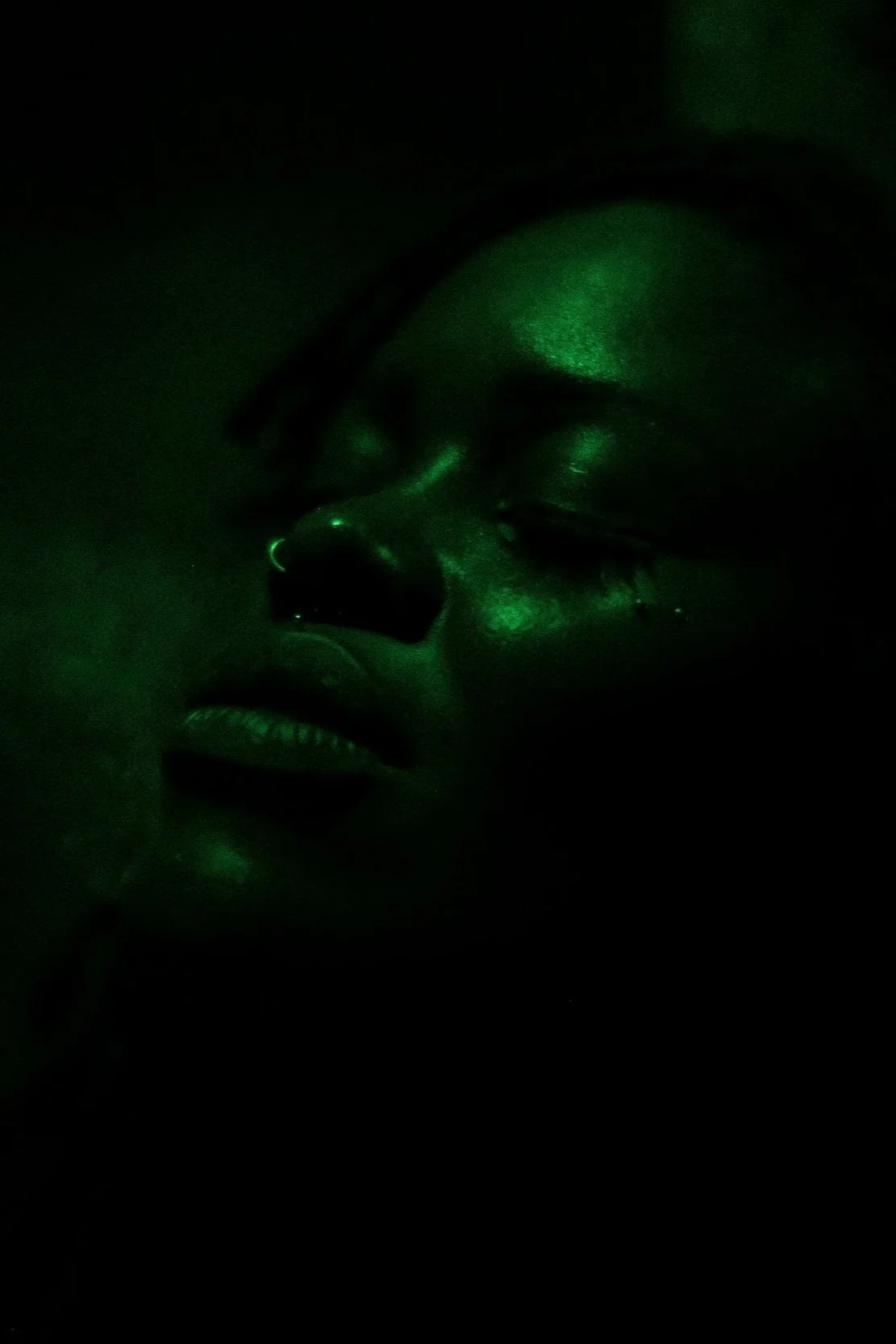 Close-up of a person's face in a dark environment with green light reflecting on skin, closed eyes, and facial features visible.