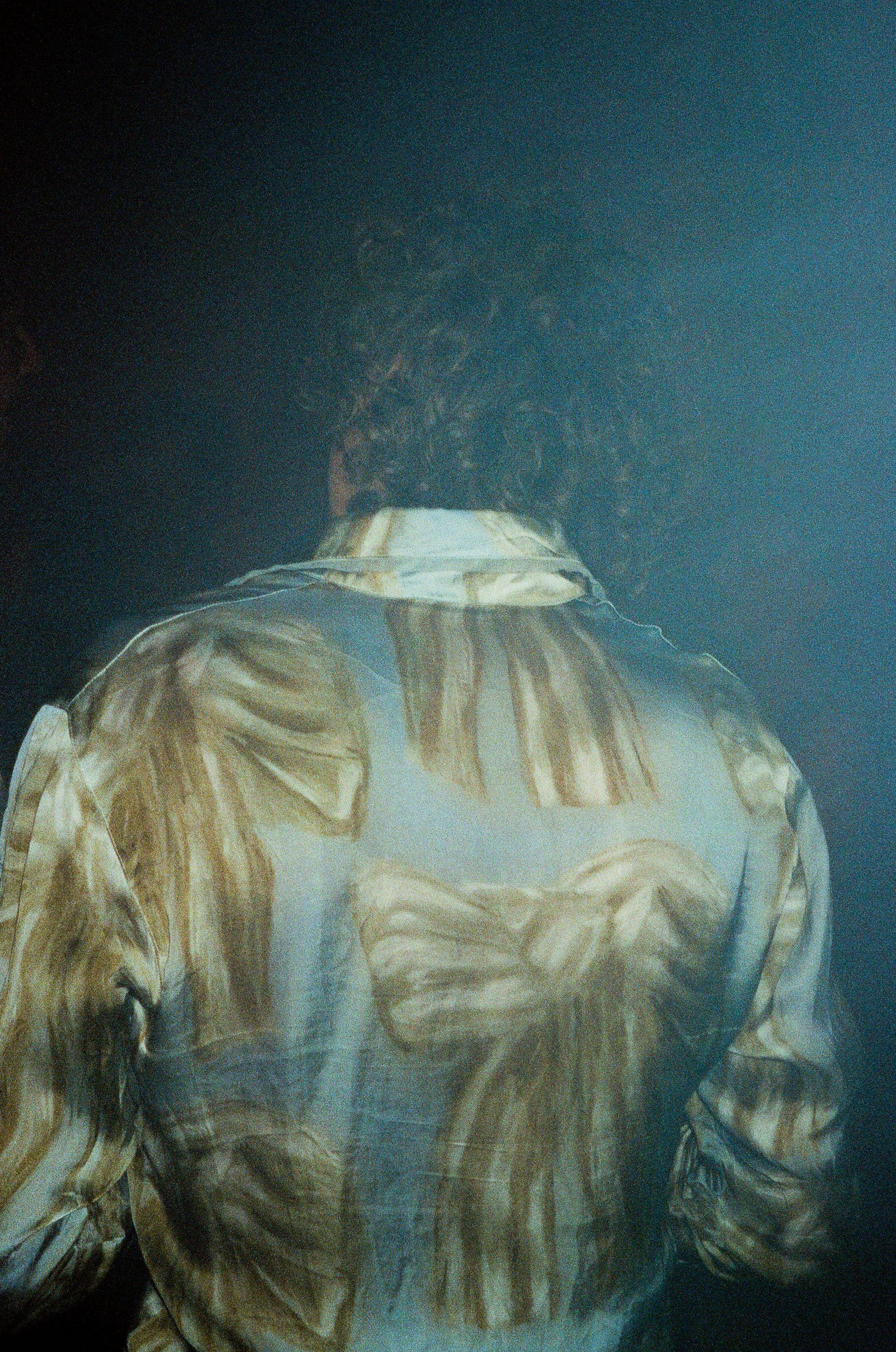 Back of a person wearing a patterned shirt with brown and tan brushstroke design, with curly hair and a dark background.