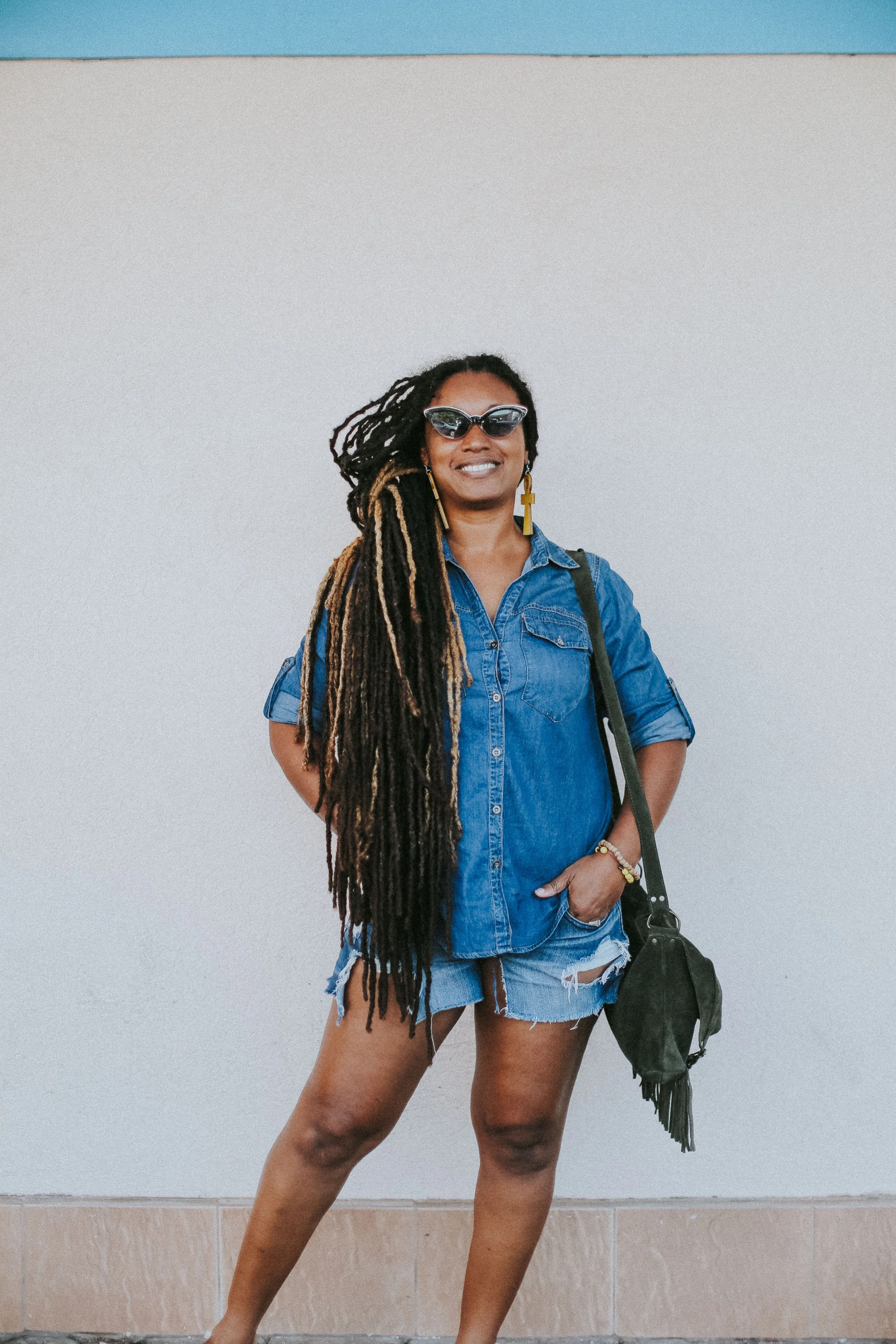 Woman with long dreadlocks, sunglasses, denim shirt, and ripped shorts standing against plain wall.