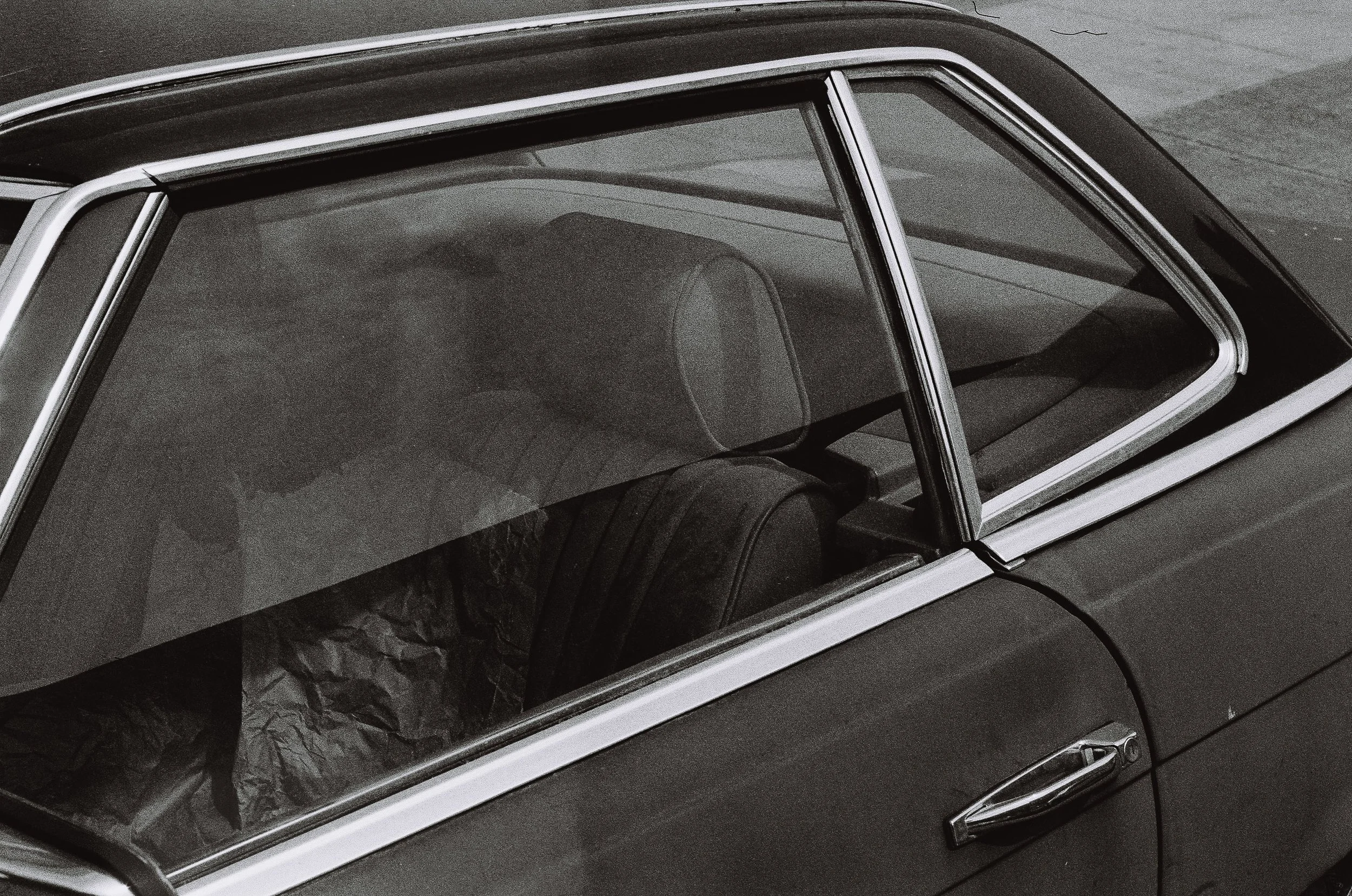 Close-up of the front passenger side of a vintage black car with a chrome door handle, showing the front windshield and interior, including a headrest and the back of the passenger seat, with some crumpled paper visible inside.
