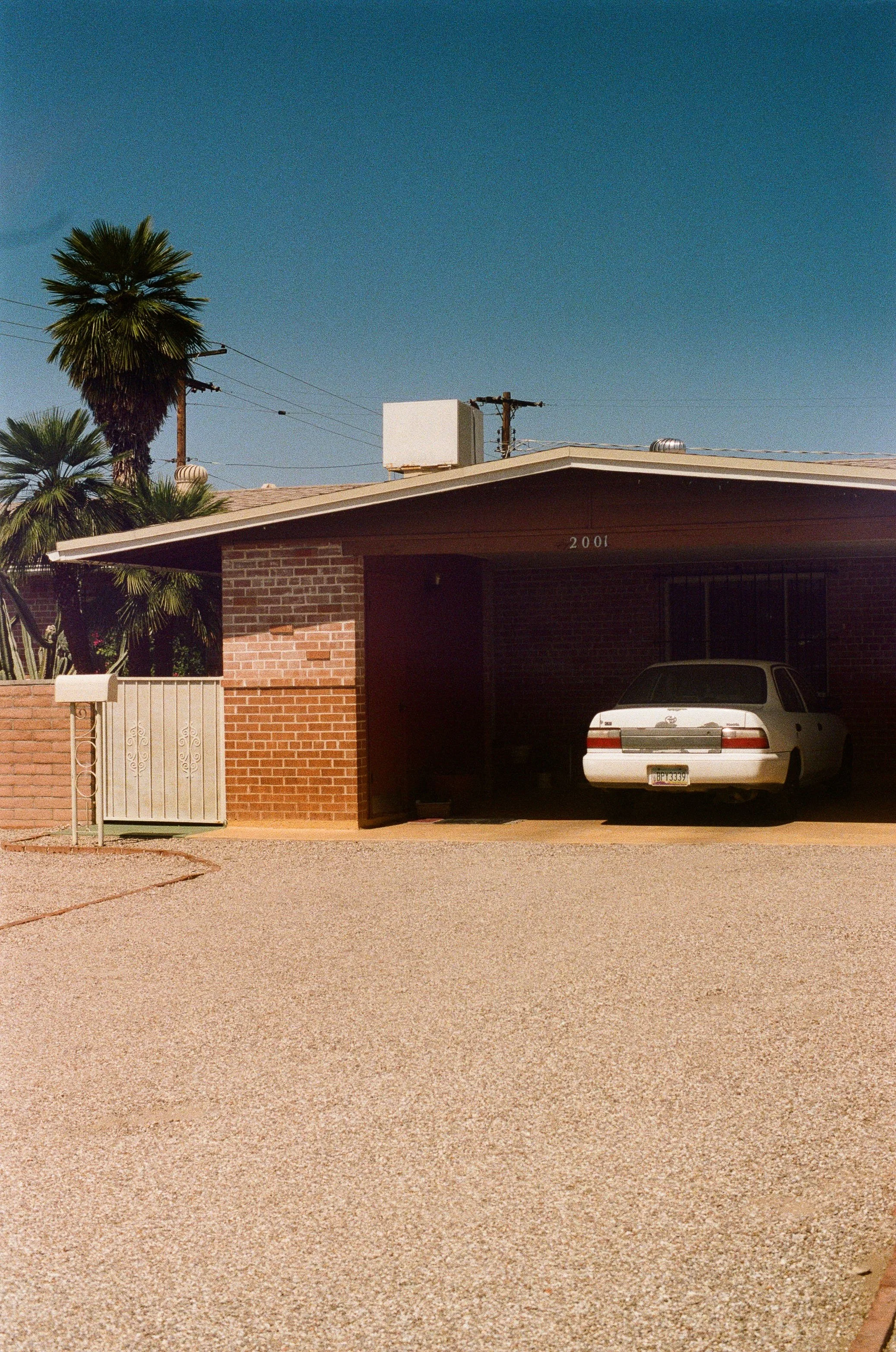 A single-story brick house with a carport, a white car parked underneath, and palm trees in the background.
