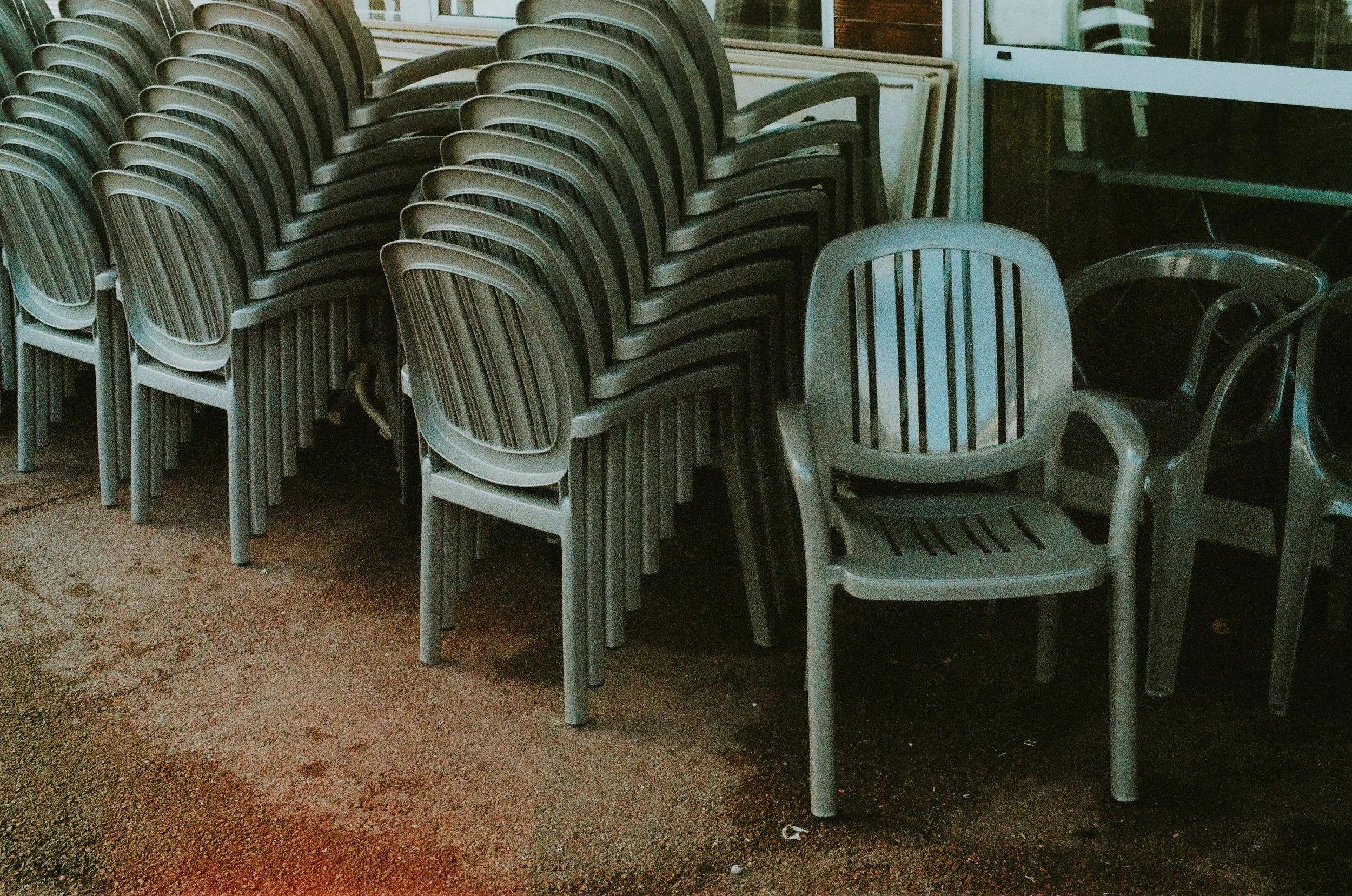 Stacked foldable outdoor chairs arranged in front of a glass door, with a few single chairs beside them, on a reddish-brown outdoor surface.