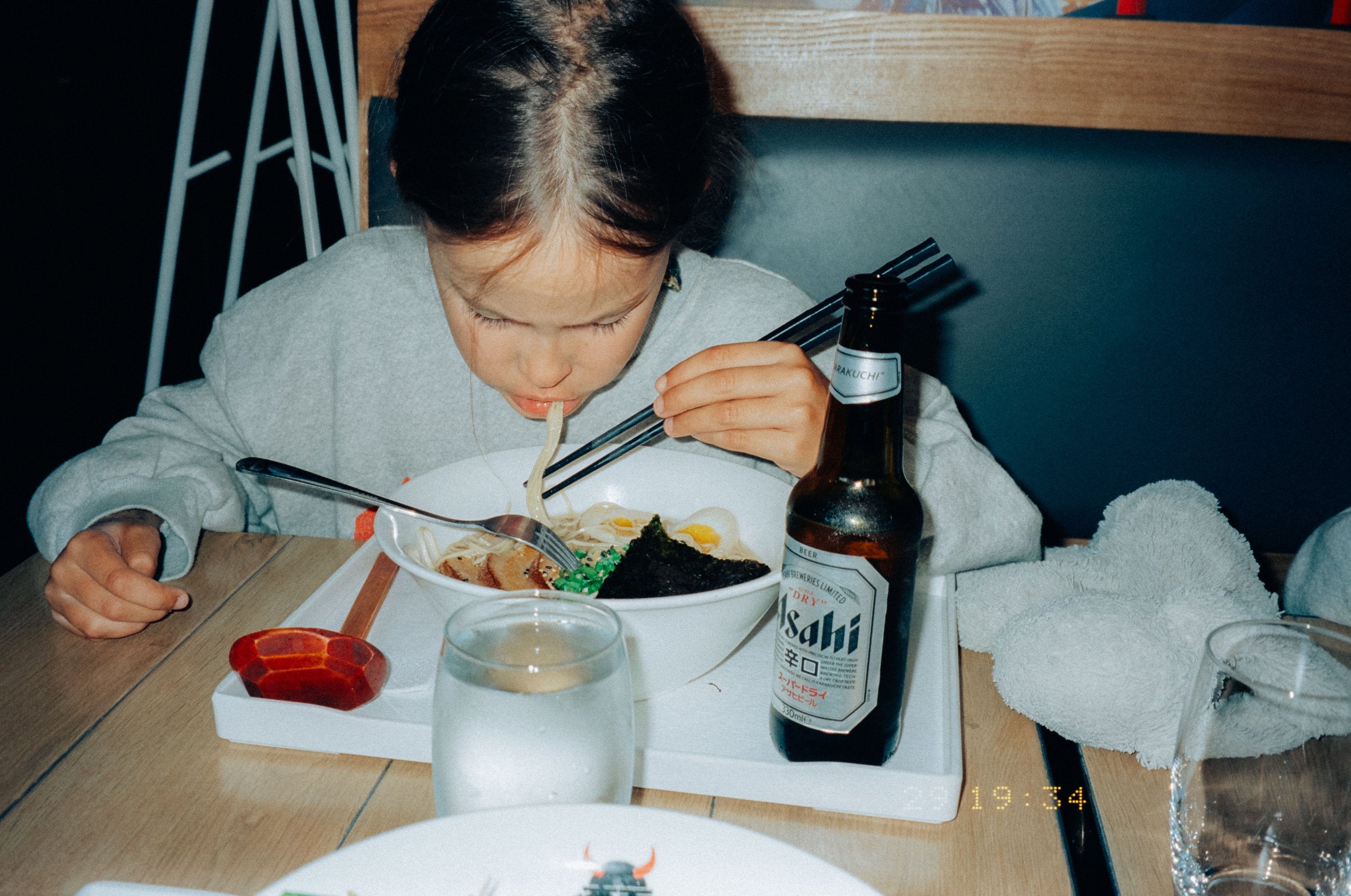 A young girl eating ramen with chopsticks at a restaurant table, with a bottle of Asahi beer, a glass of water, a folded towel, and plates in the background.