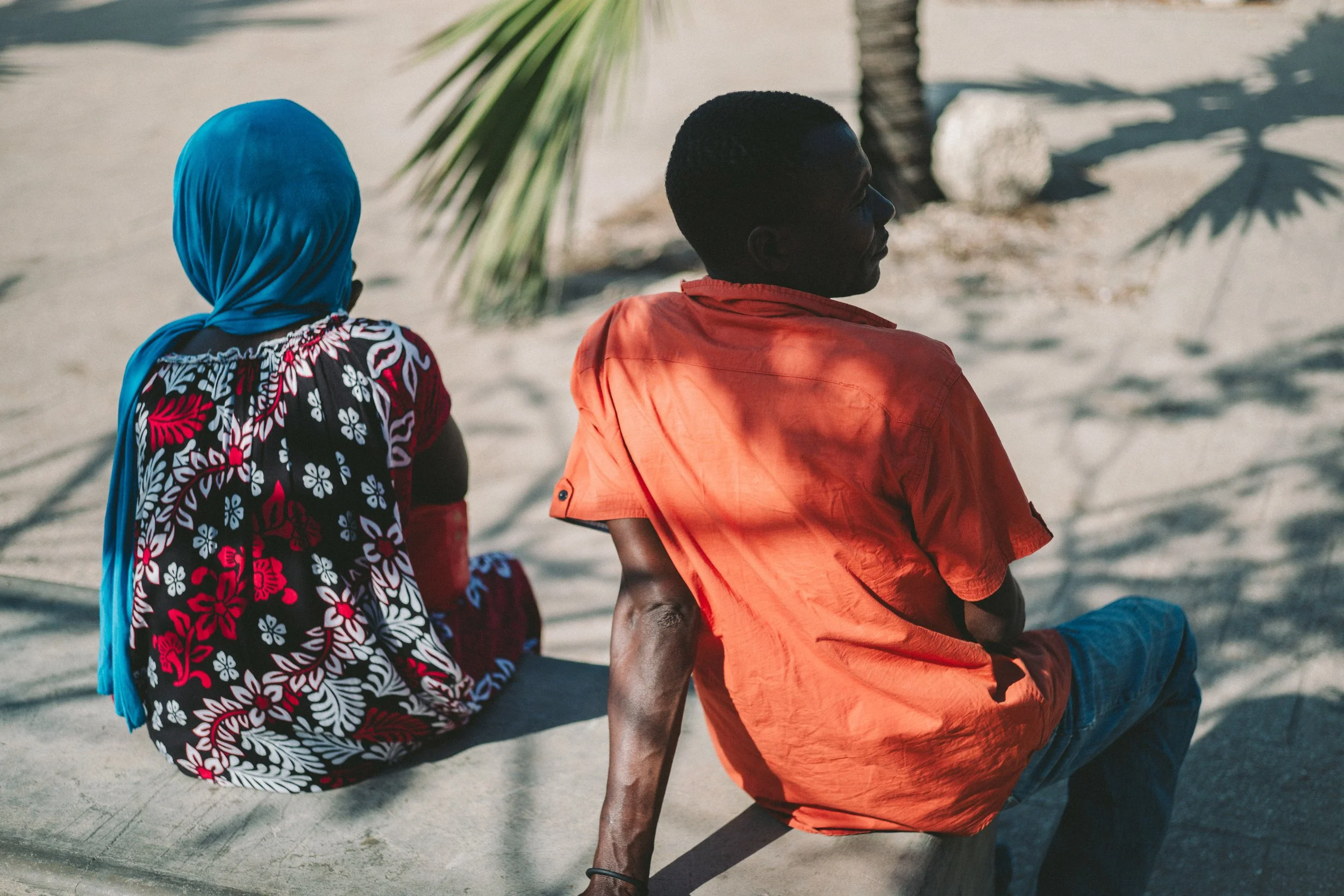 A woman wearing a blue headscarf and a dress with a black, red, and white floral pattern sits on a concrete surface next to a man in an orange shirt and blue jeans. They are outdoors near a palm tree and casting shadows on the ground.