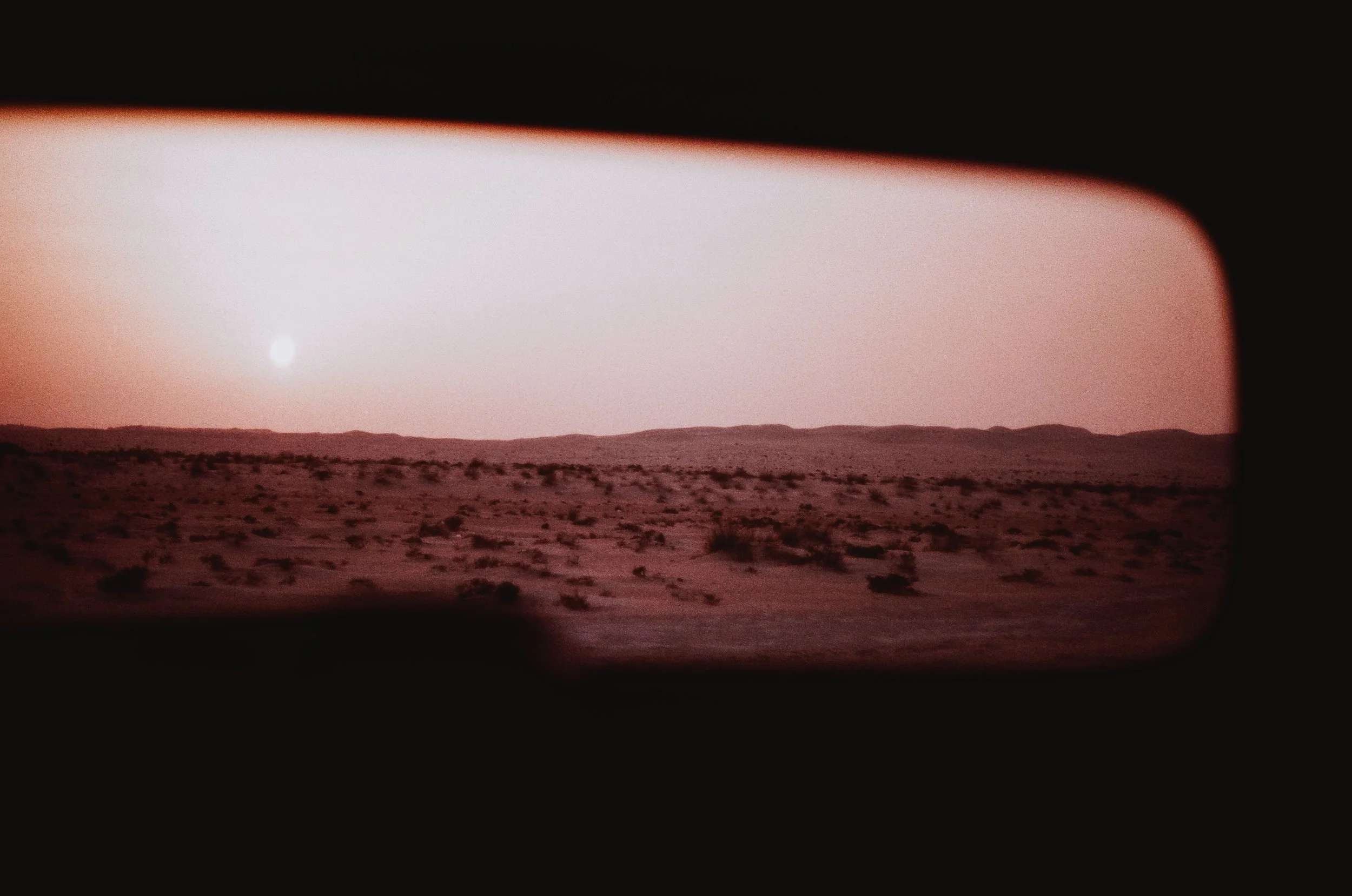 A desert landscape viewed through a car's rearview mirror, with the sun setting near the horizon and pinkish sky.