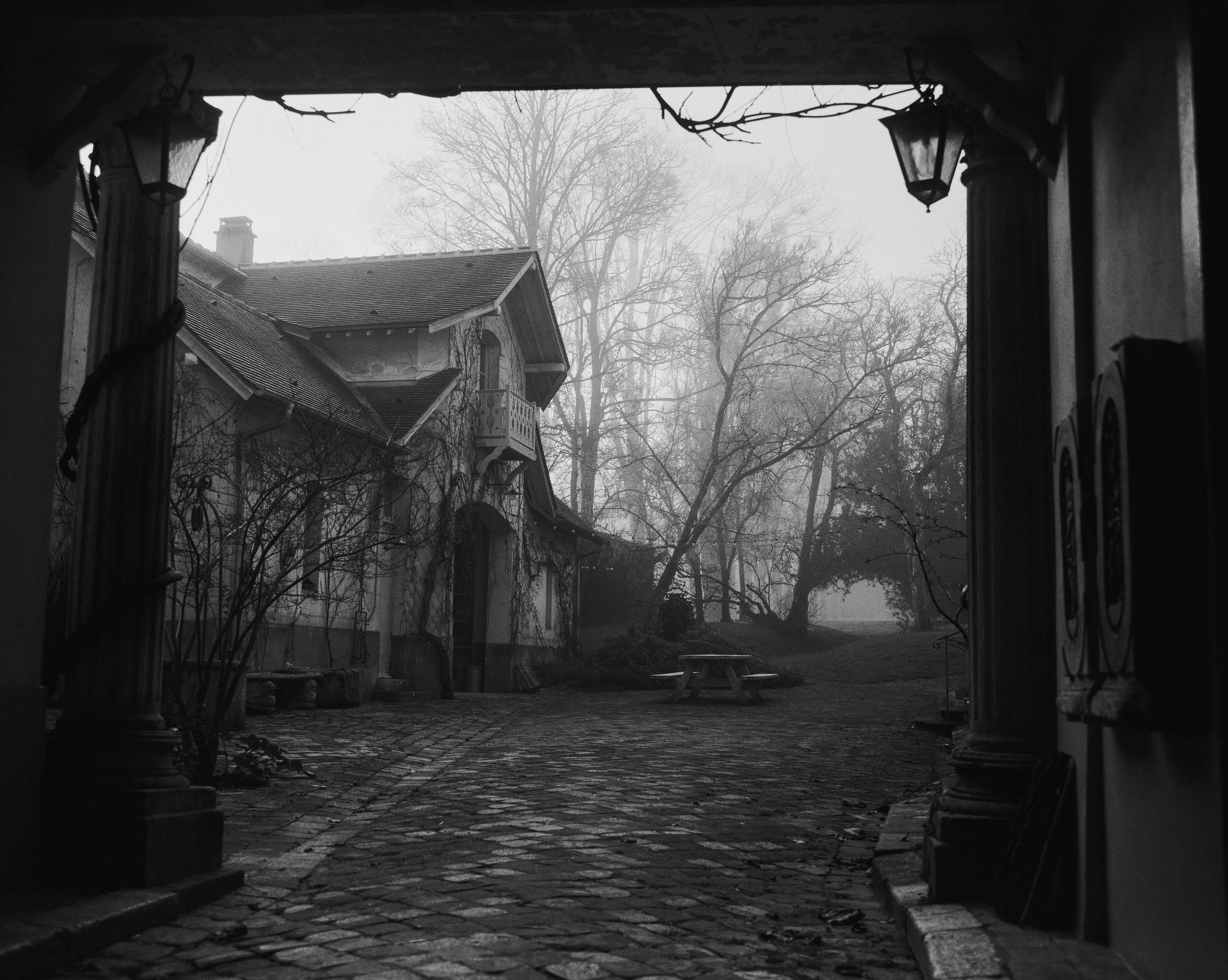 Black and white image of a foggy courtyard viewed from underneath an archway with lanterns. There are leafless trees, an old house with a small balcony, a picnic table, and cobblestone ground.