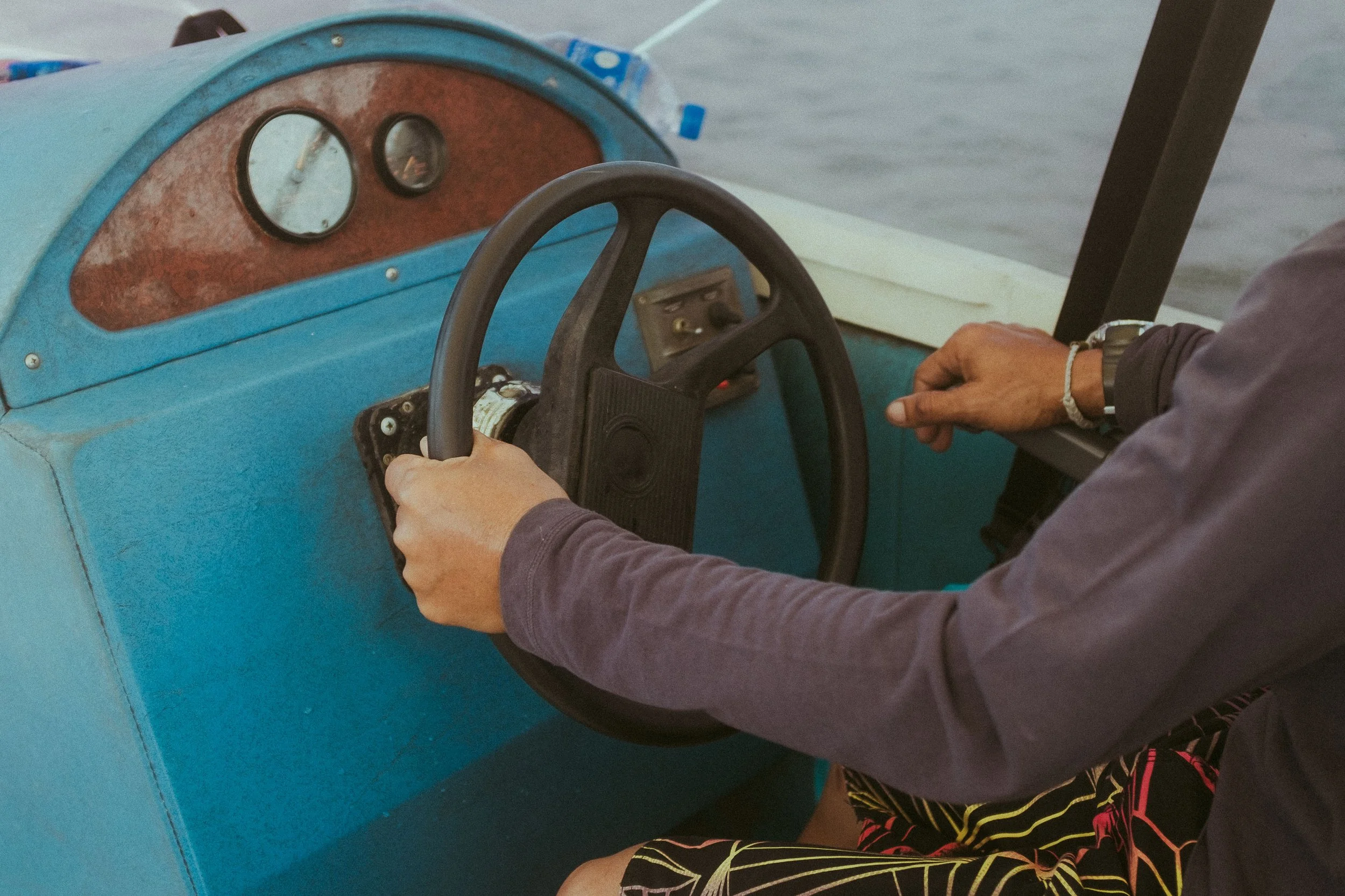 Person steering a small boat with a black steering wheel on a blue dashboard, water visible in the background.