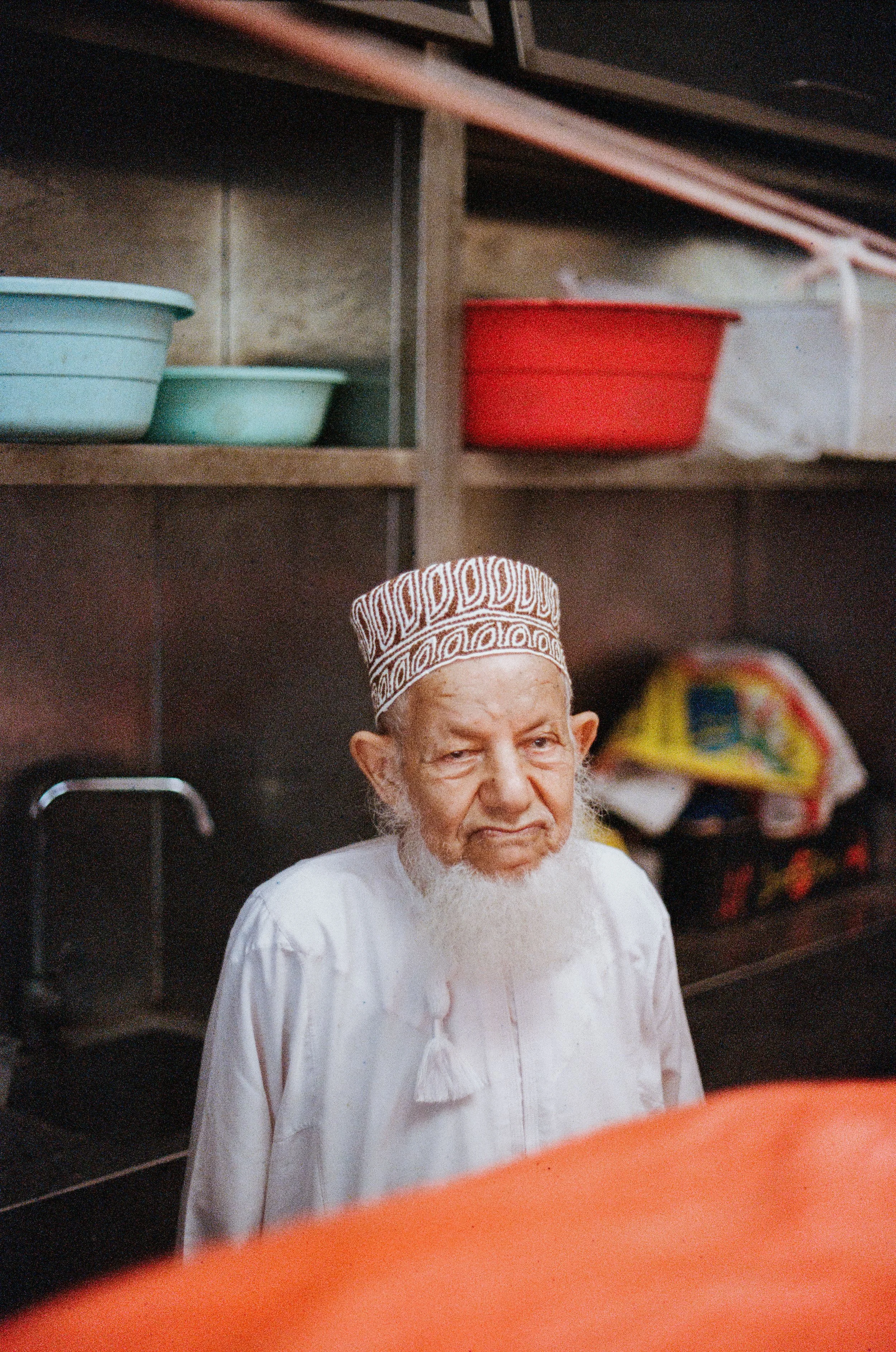 An elderly man with a white beard and traditional cap standing in a kitchen with shelves of colorful bowls and plastic bags in the background.