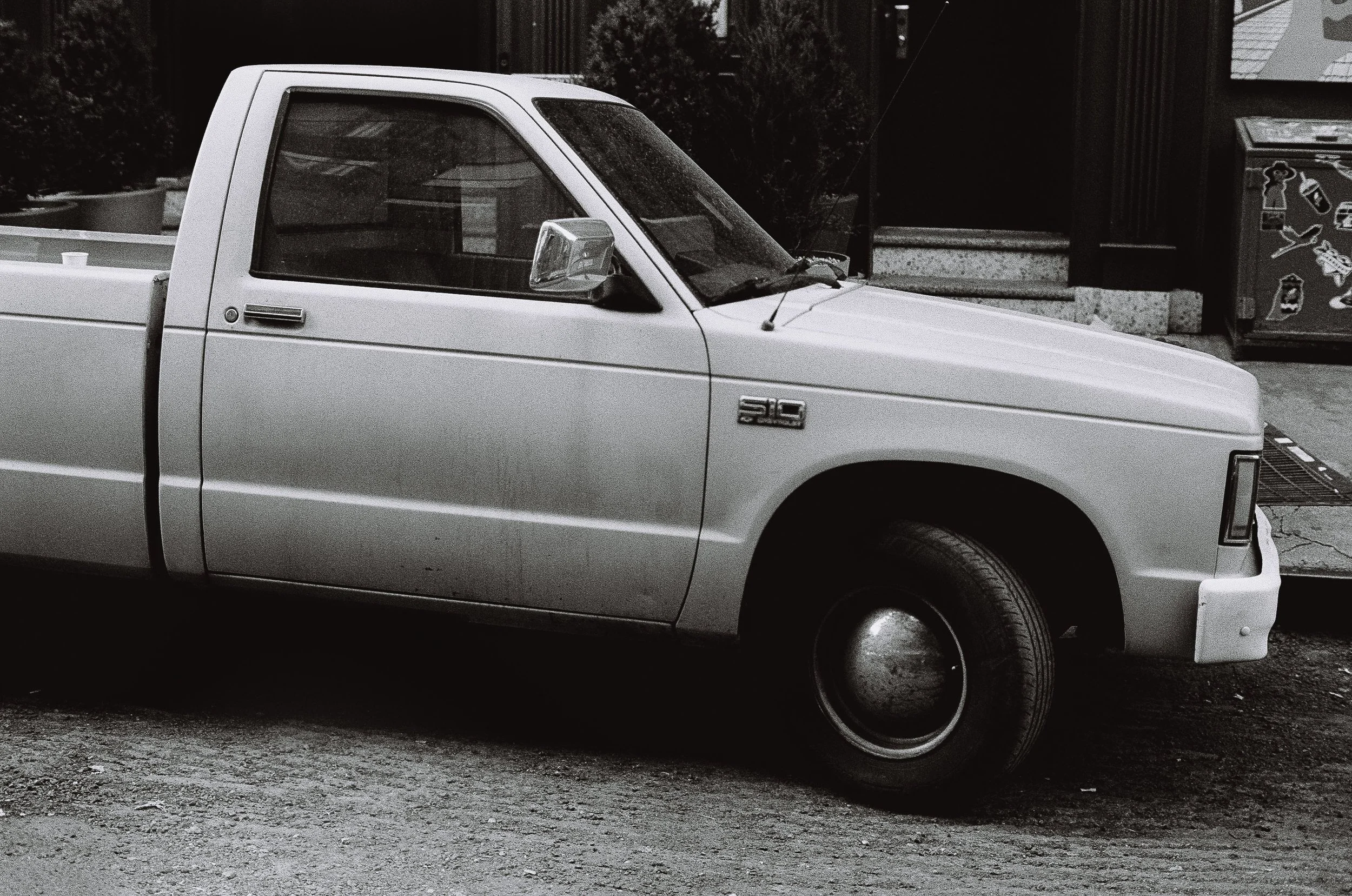 A black and white photo of a vintage pickup truck parked on the street, with a side view showing the front and part of the side, including the driver's side door and mirror.