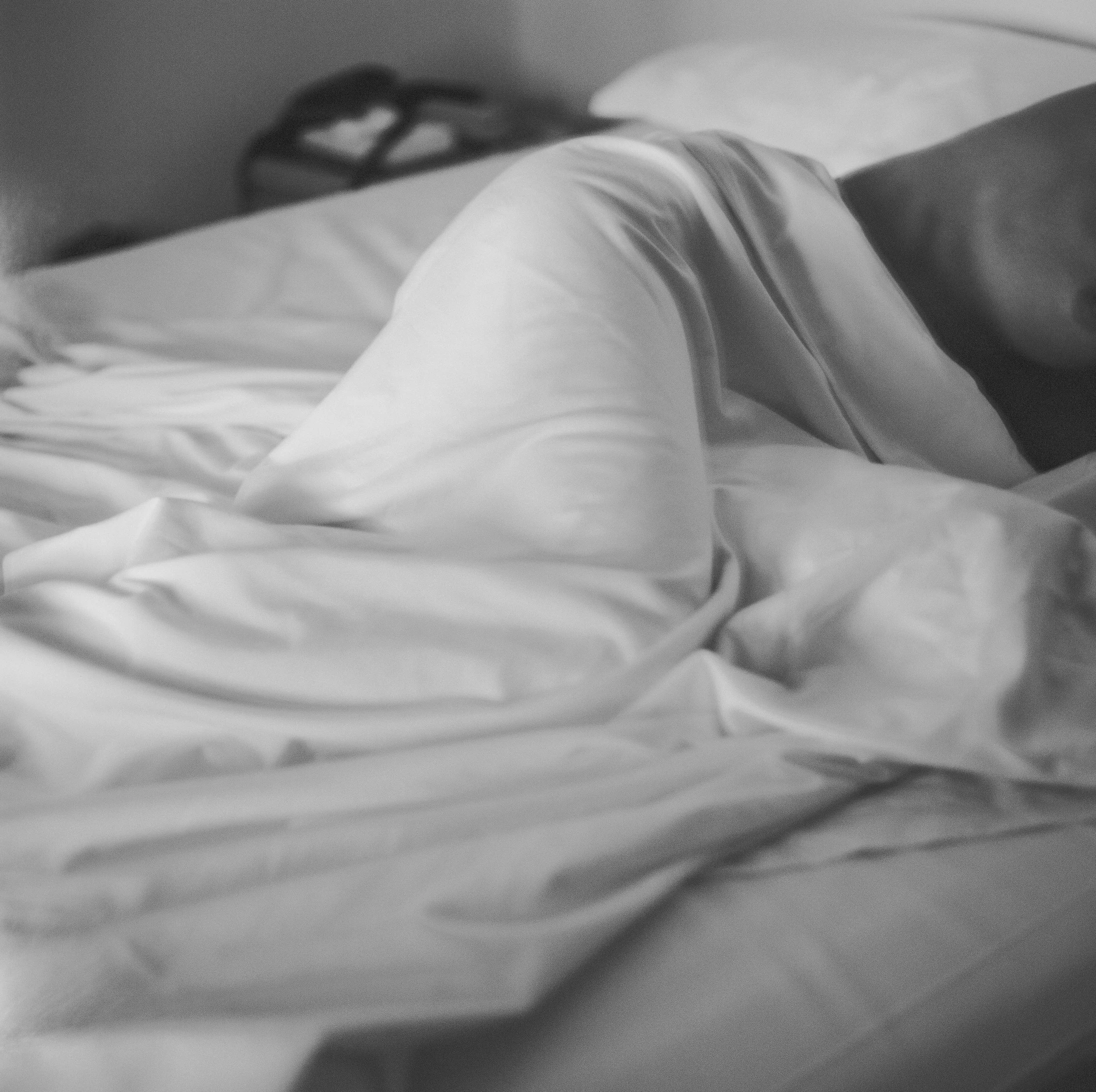 A person lying on a bed with a white sheet, wearing a white shirt, and resting with eyes closed.