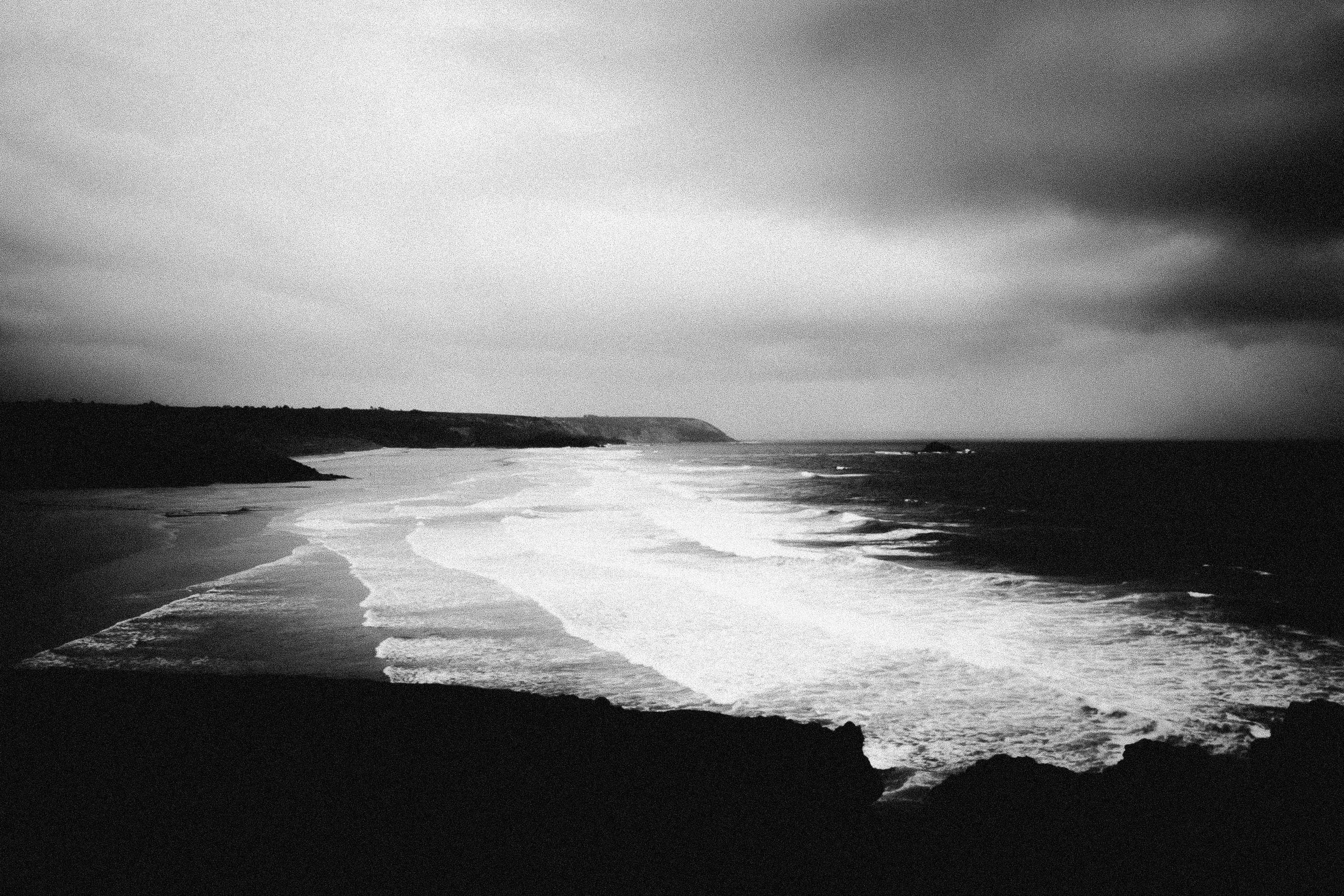Black and white photograph of a rugged coastline with waves crashing onto the shore, dark clouds overhead, and cliffs in the distance.