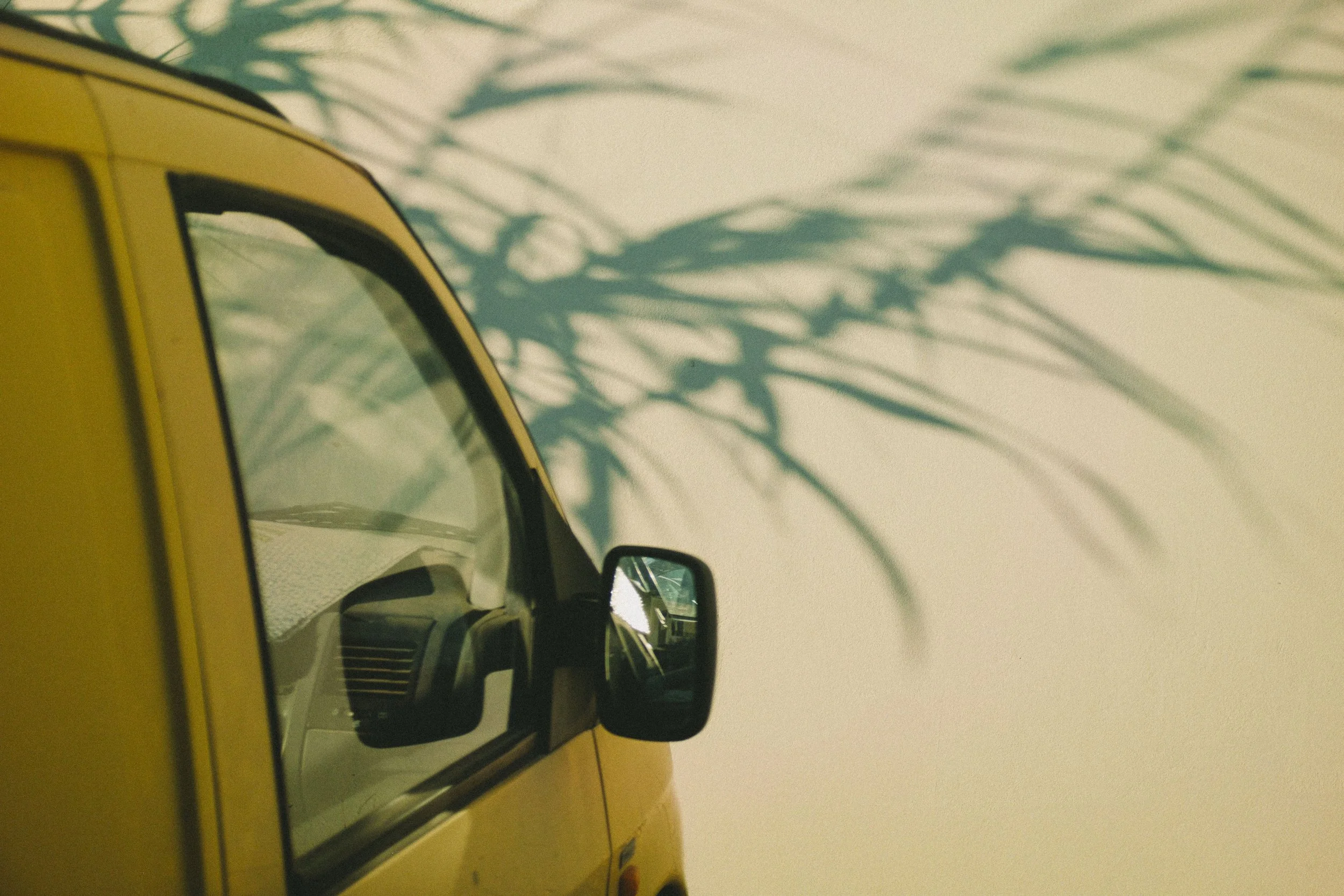 Part of a yellow vehicle, possibly a van, with a side mirror visible. The vehicle is casting shadow of leaves or branches on the wall behind it.