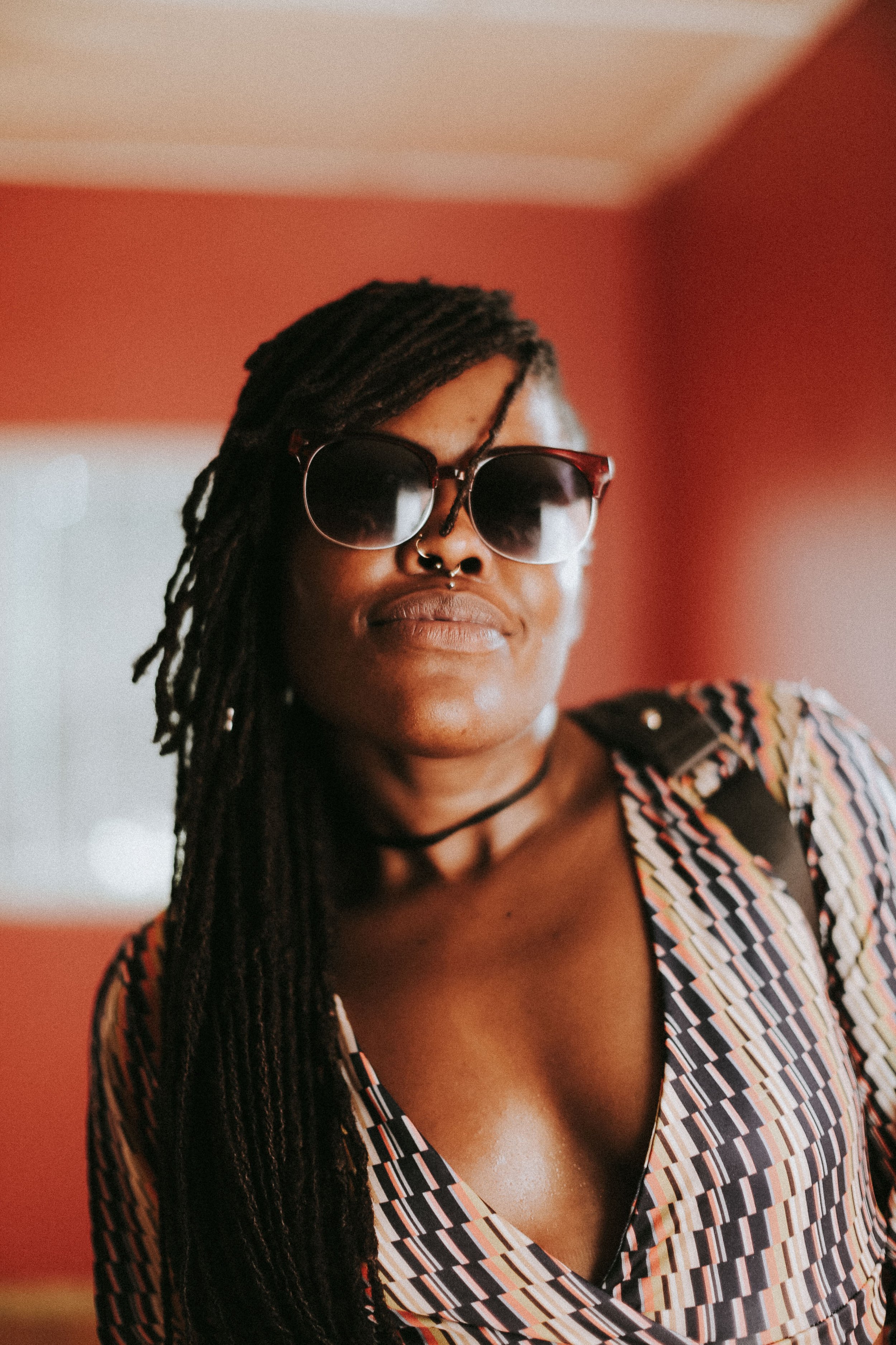 A woman with long dreadlocks wearing dark sunglasses, a septum piercing, a choker, and a patterned top, looking confidently at the camera against a warm-colored background.