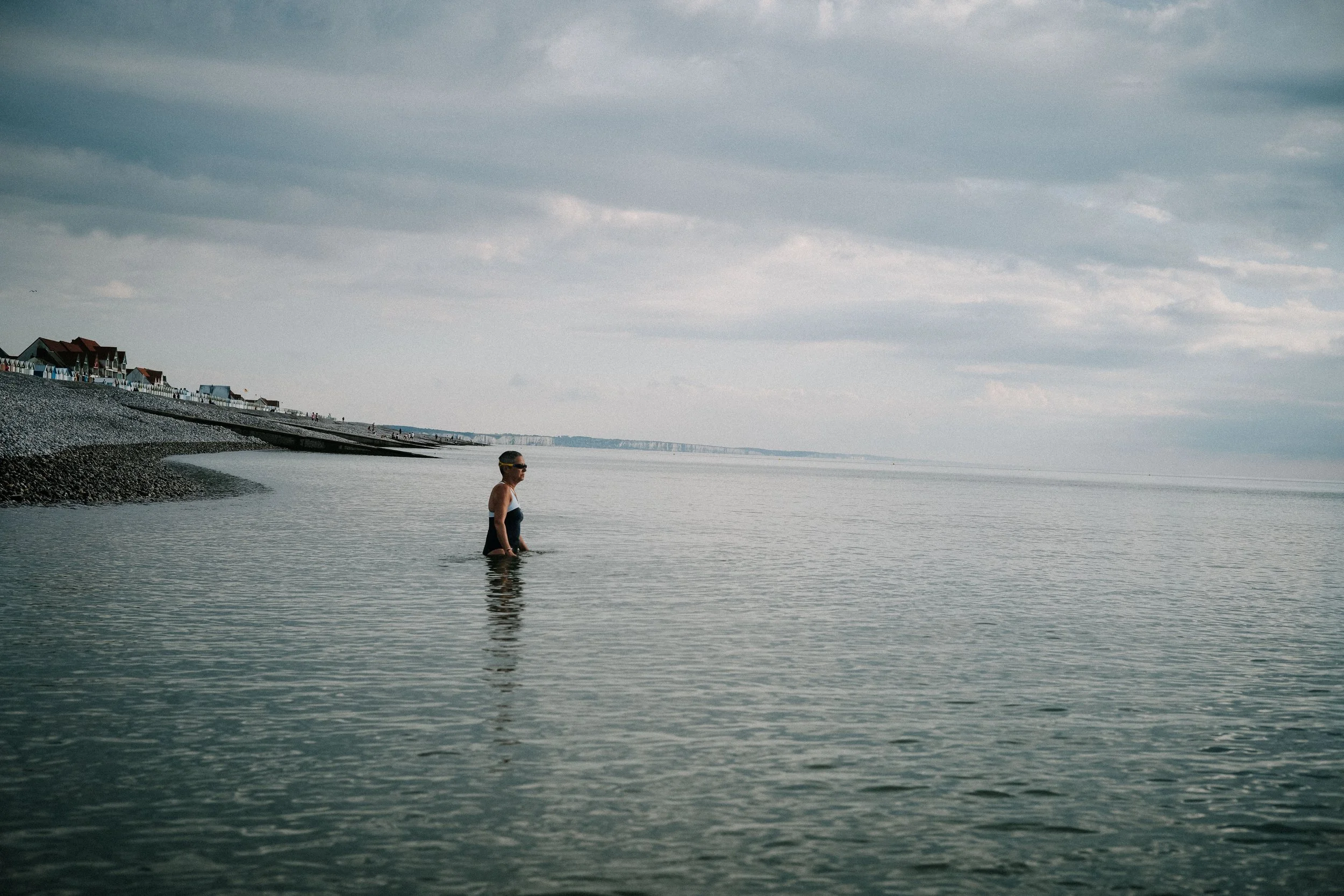 A woman stands in shallow sea water near a pebble beach on a cloudy day, with houses visible on the shore in the background.