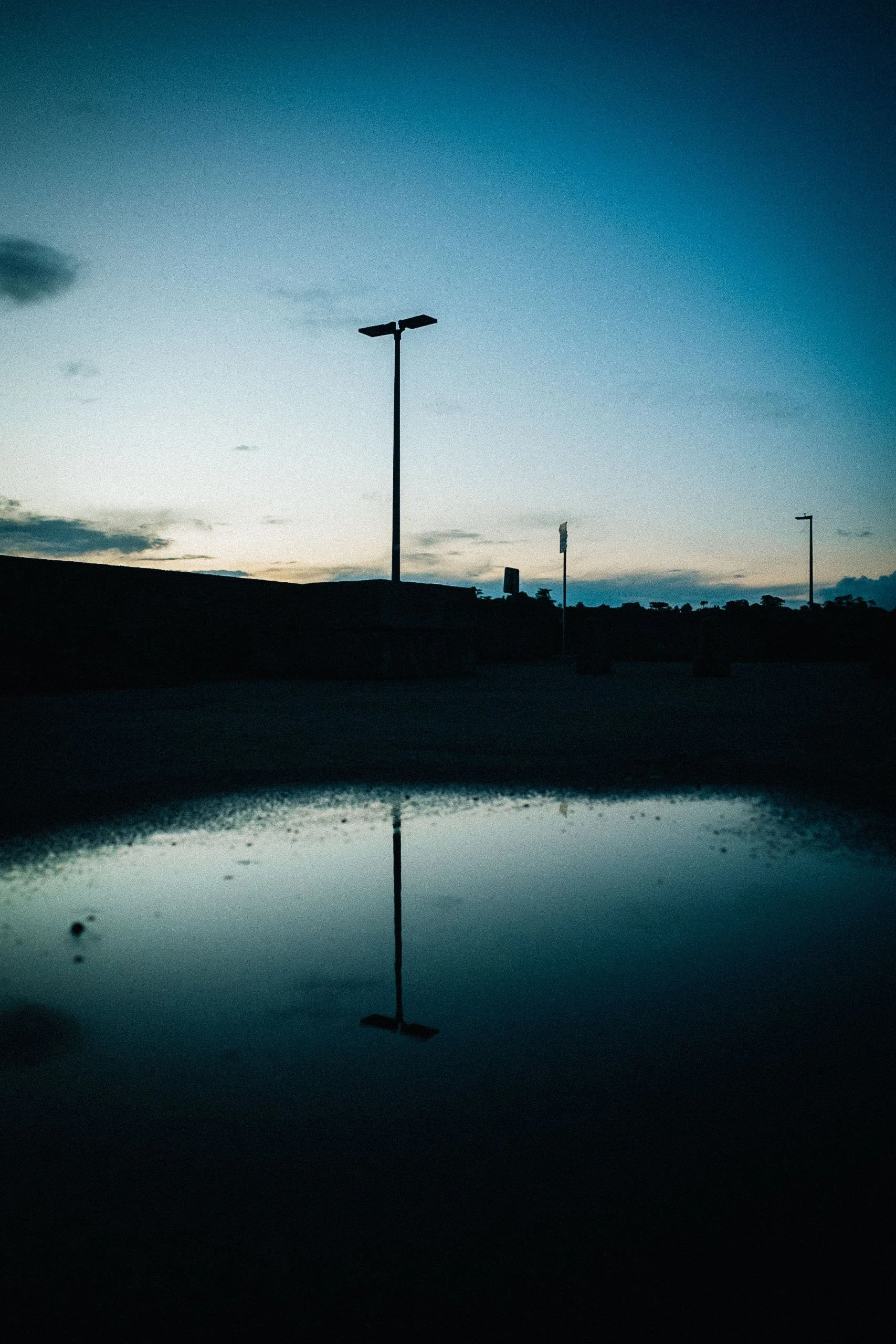 Silhouette of streetlights and a sign against a sunset sky with a reflection in a dark puddle.