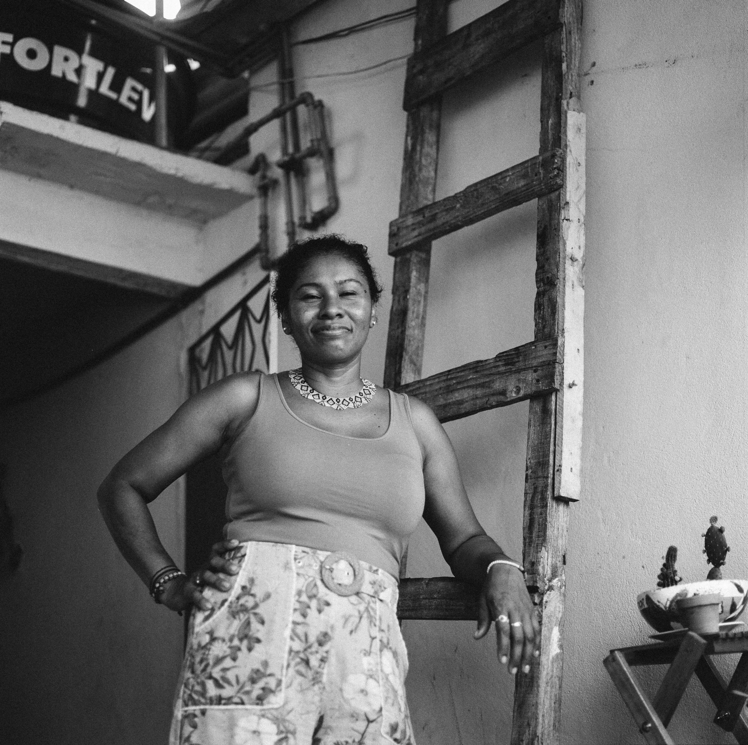 A smiling woman standing indoors, wearing a sleeveless top, floral skirt, and jewelry, with her hand resting on a wooden ladder next to her.