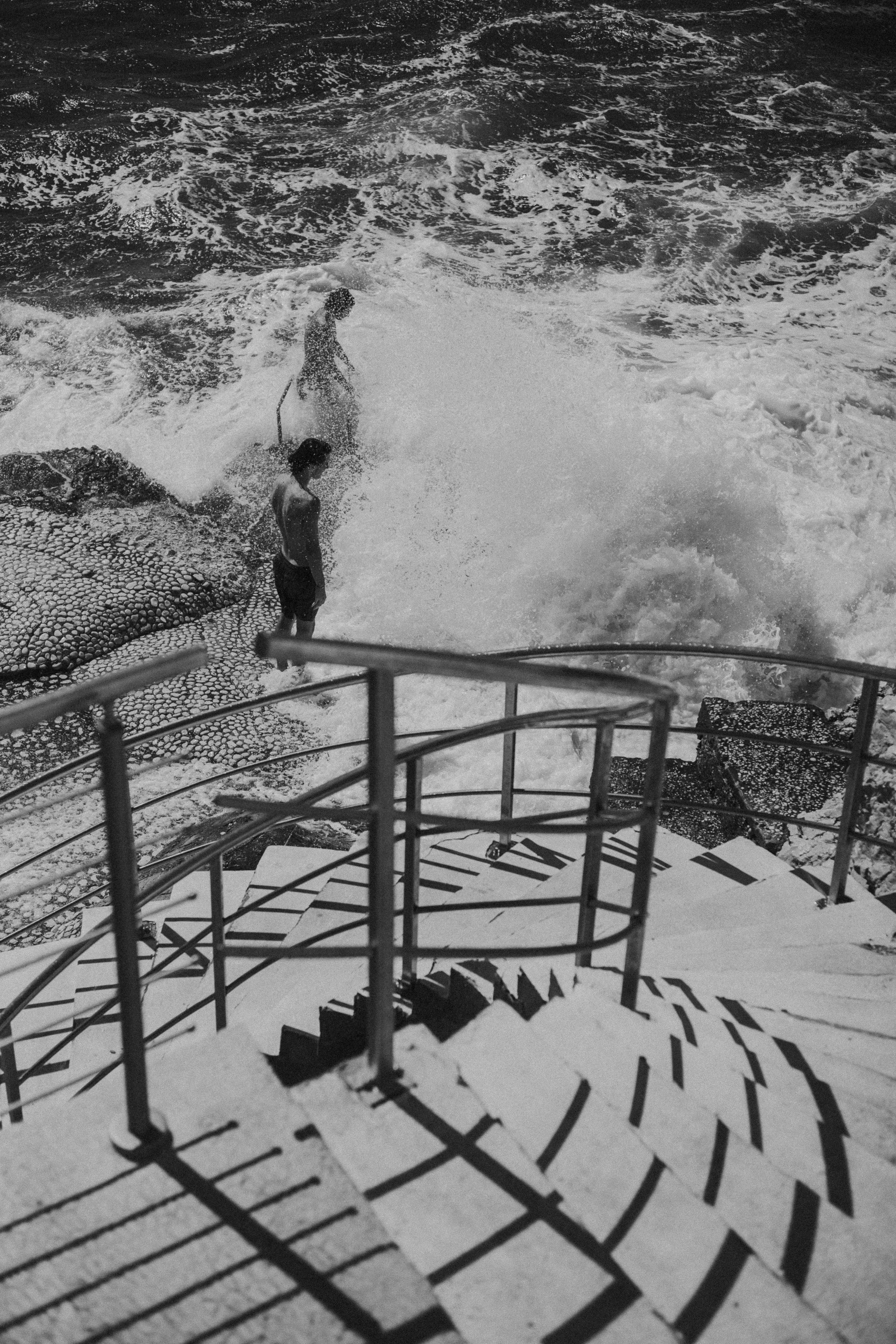 Black and white photo of two people near the ocean, one standing on rocks and the other in the water, with steps and a railing in the foreground.