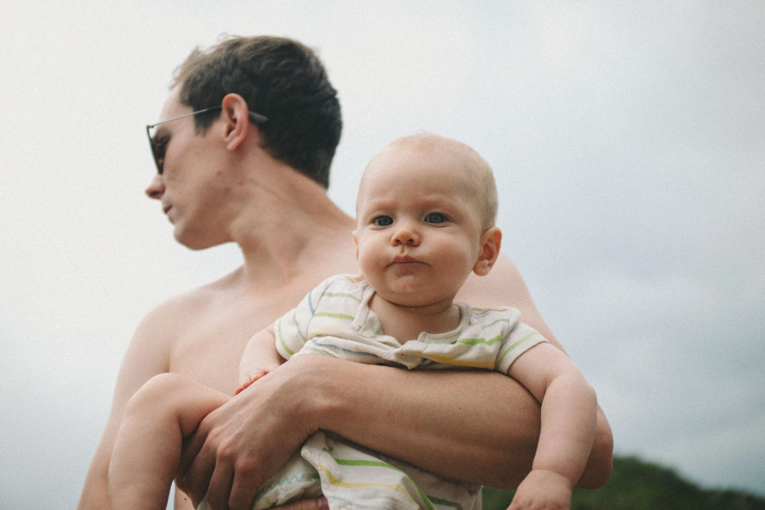 A shirtless man holding a young child outdoors against a cloudy sky.
