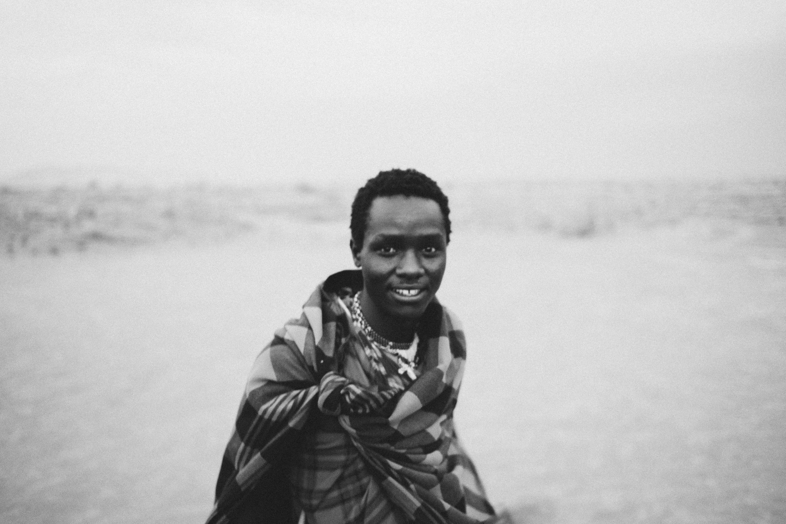 A person with short curly hair standing on a beach, wrapped in a patterned cloth, smiling at the camera with the ocean in the background.