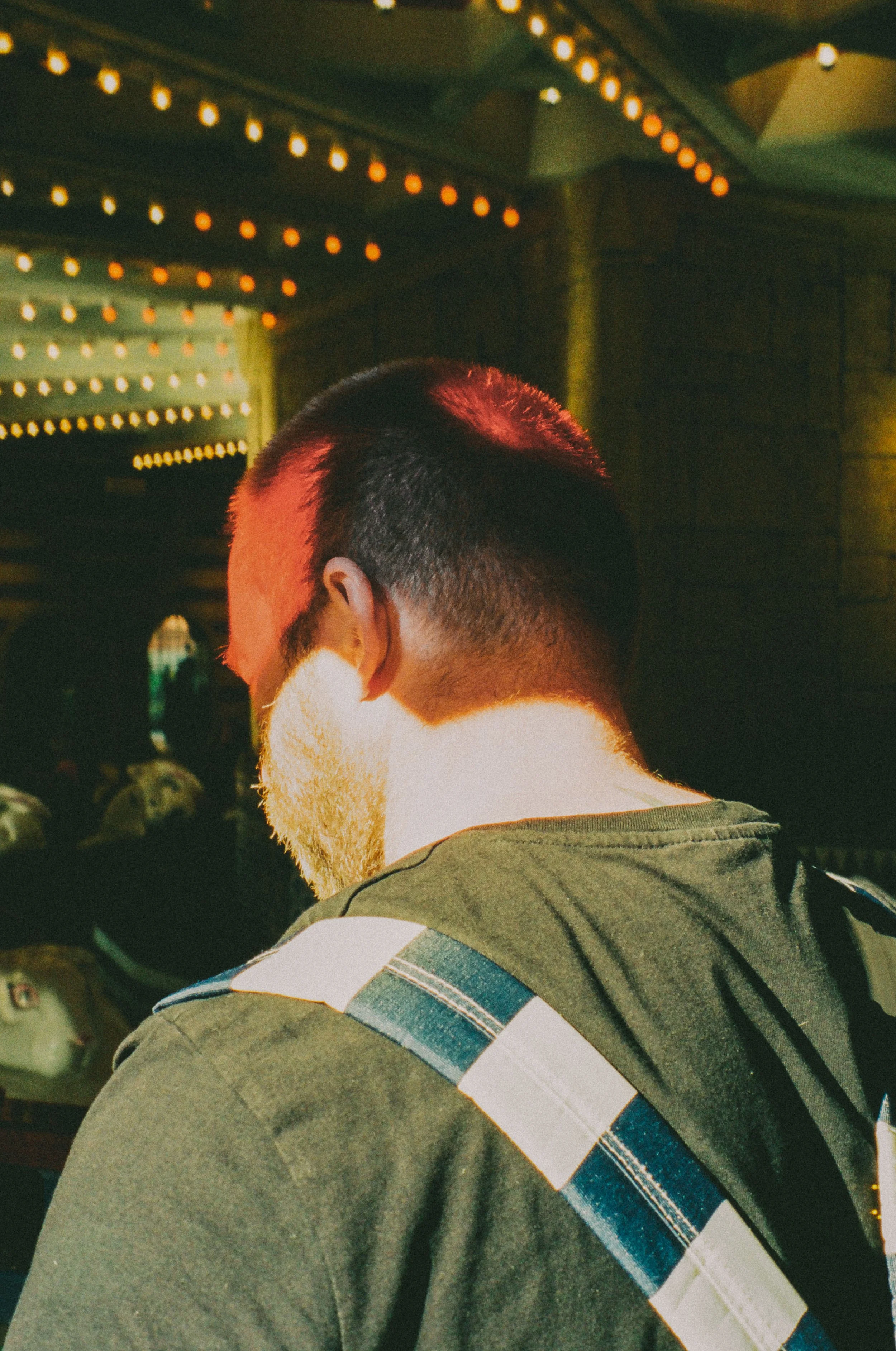 Back of a man with short dark hair and a light beard, wearing a dark green shirt with a green, white, and blue striped strap across the shoulder, in a dimly lit setting with string lights overhead.