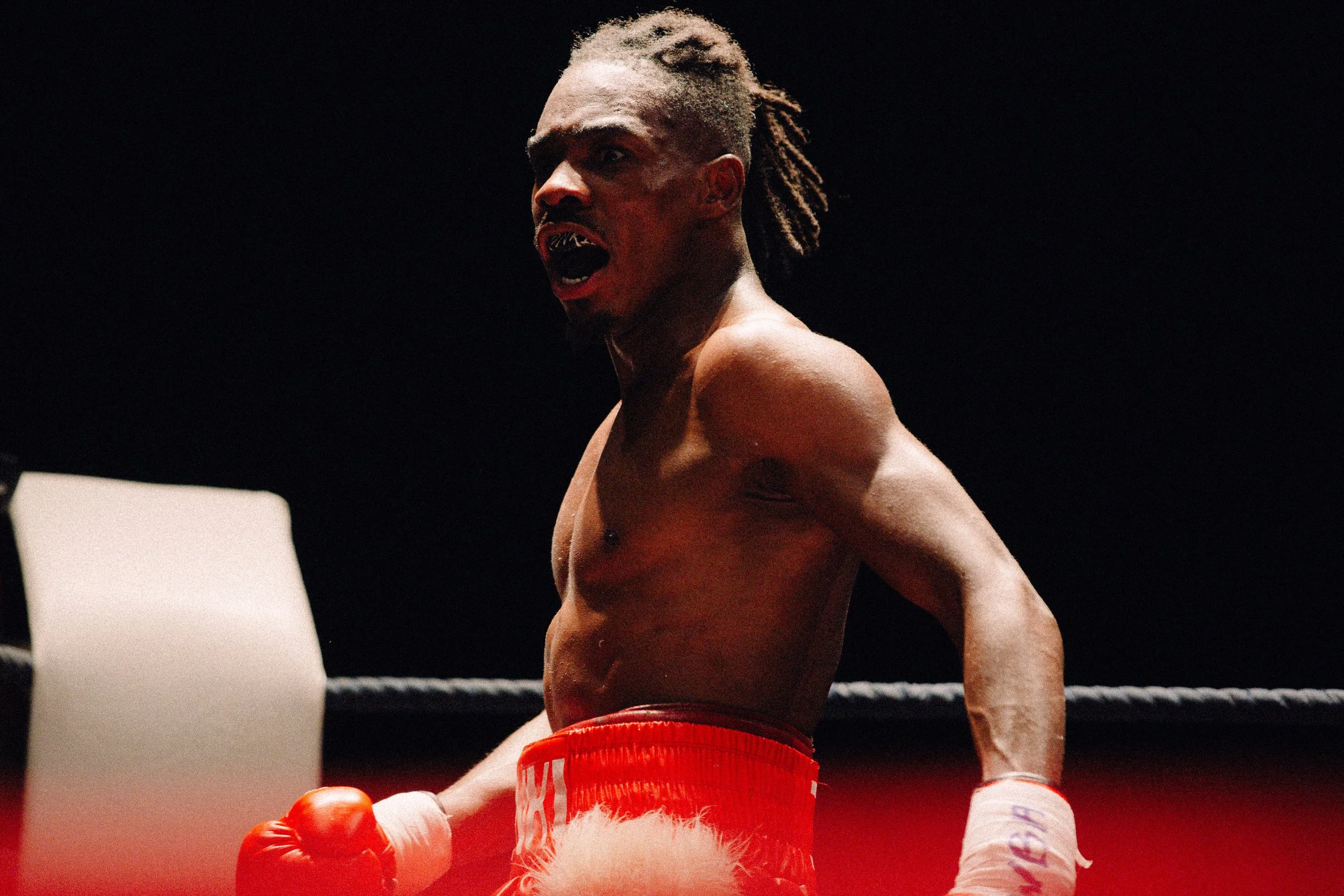 A shirtless male boxer with dreadlocks wearing red boxing trunks and gloves, standing inside a boxing ring with a black background, looking intensely with his mouth open.