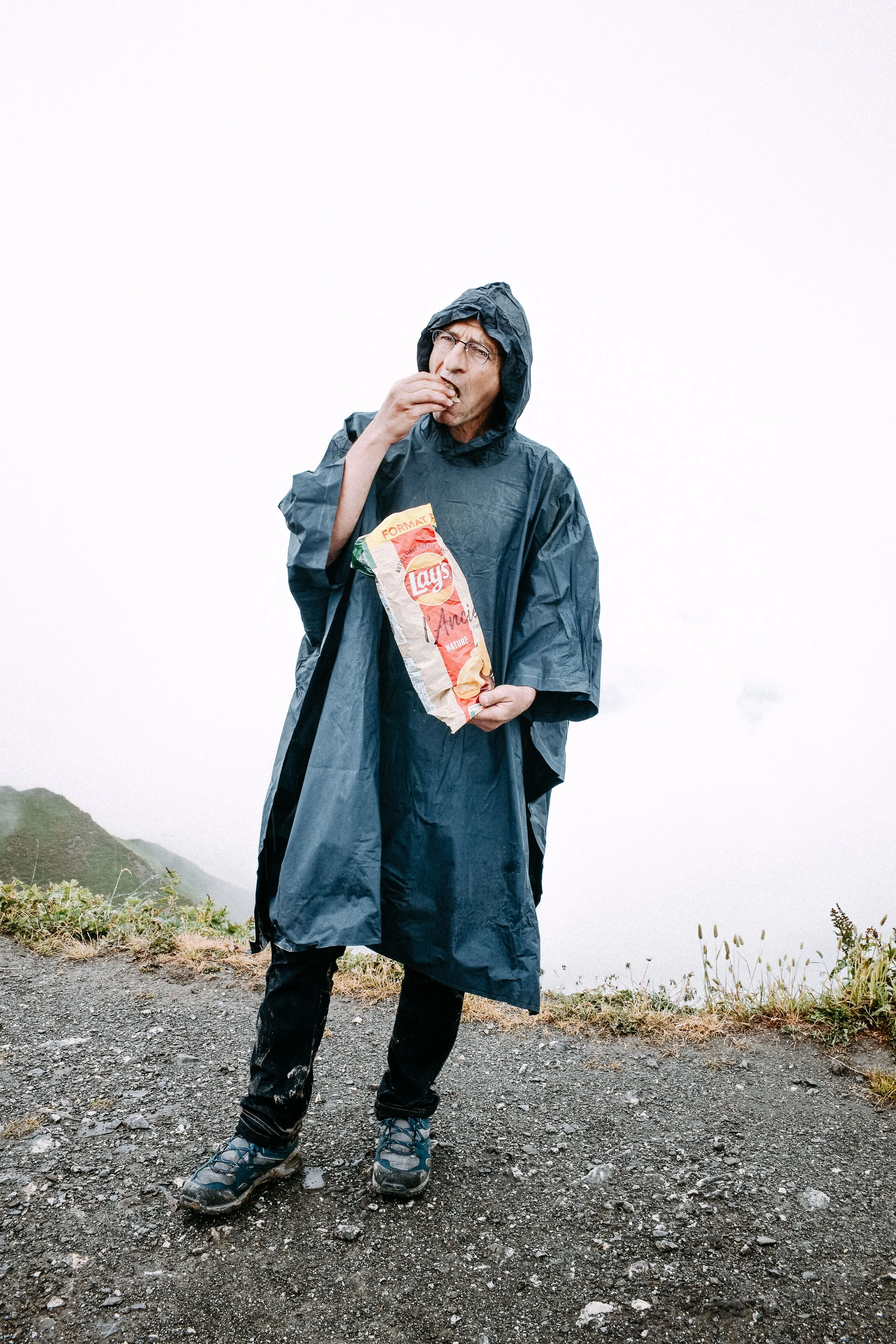 A man standing outdoors on a cloudy, foggy mountain trail, wearing a raincoat and hat, eating potato chips from a Lay's bag.