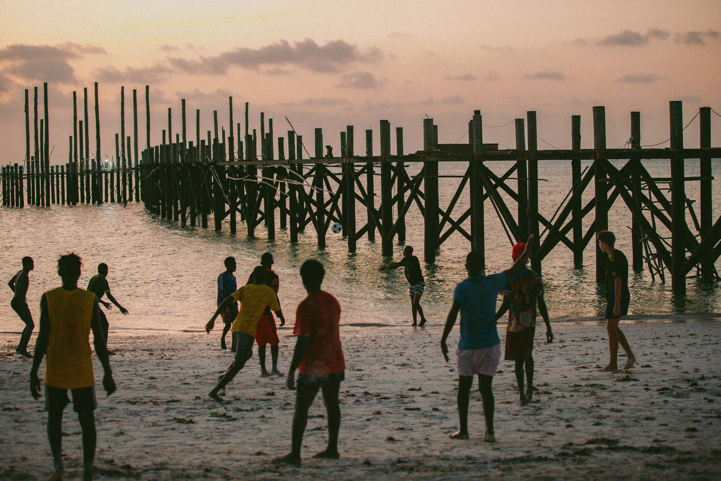 Children playing soccer on the beach at sunset near a wooden pier.