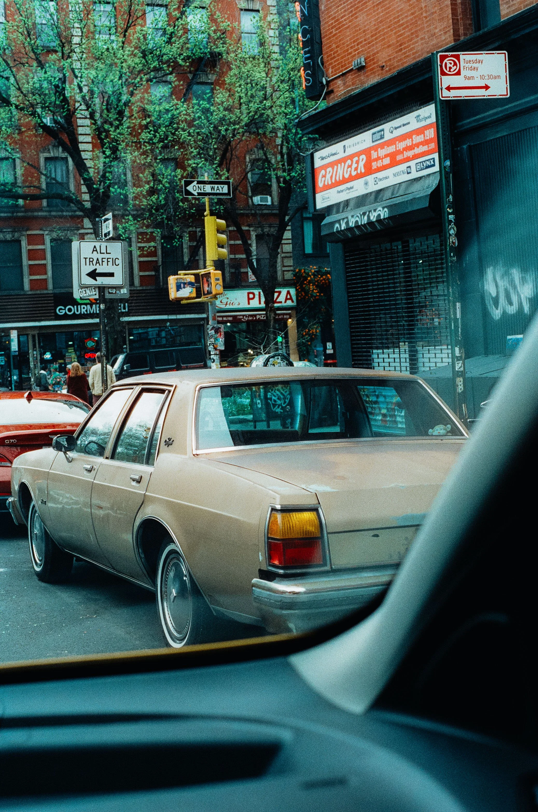 A beige vintage car parked on a city street with trees, storefronts, and signs in the background.