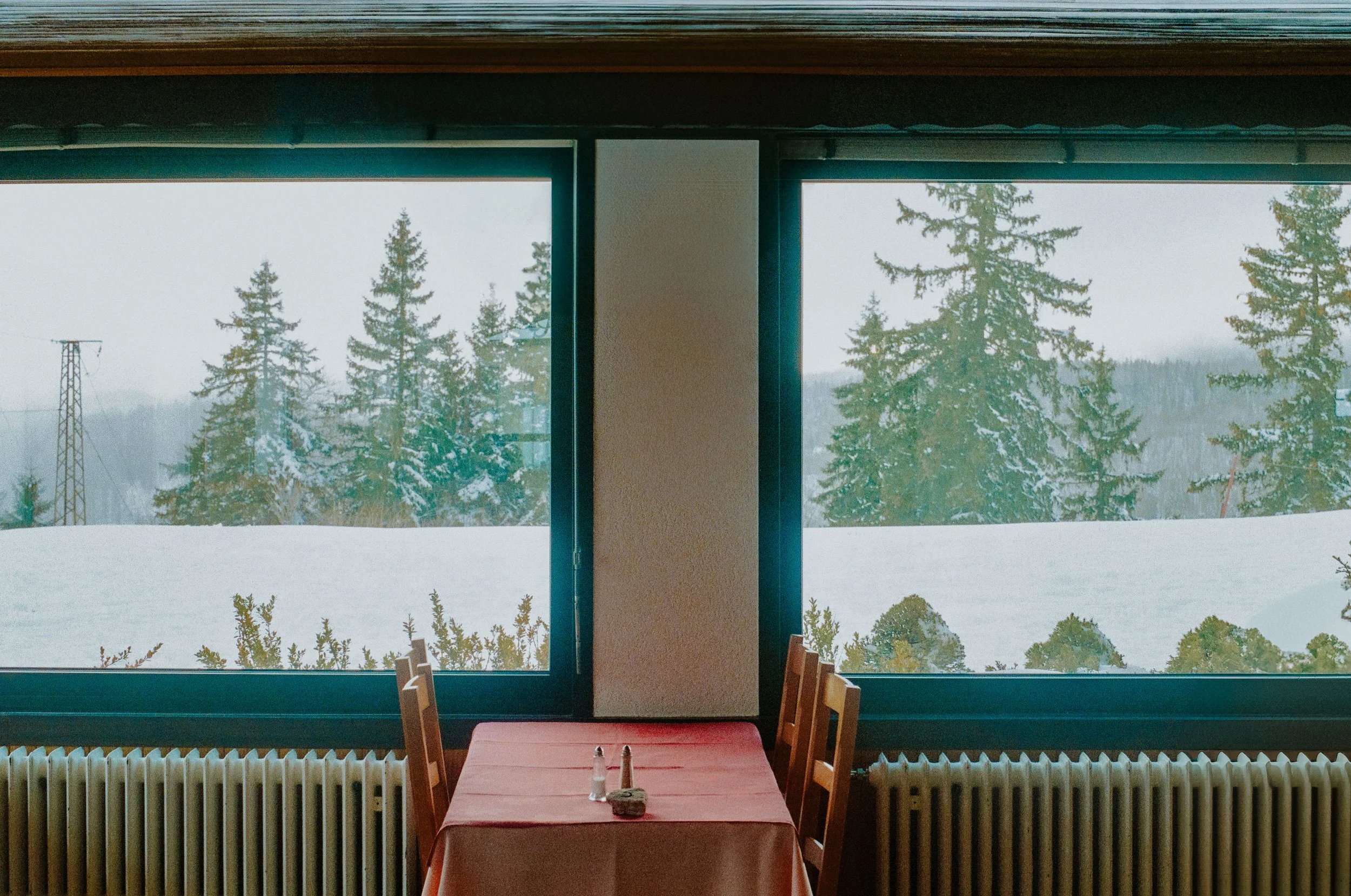 Empty restaurant with a table set for two, located by large windows showing snowy trees and landscape outside.