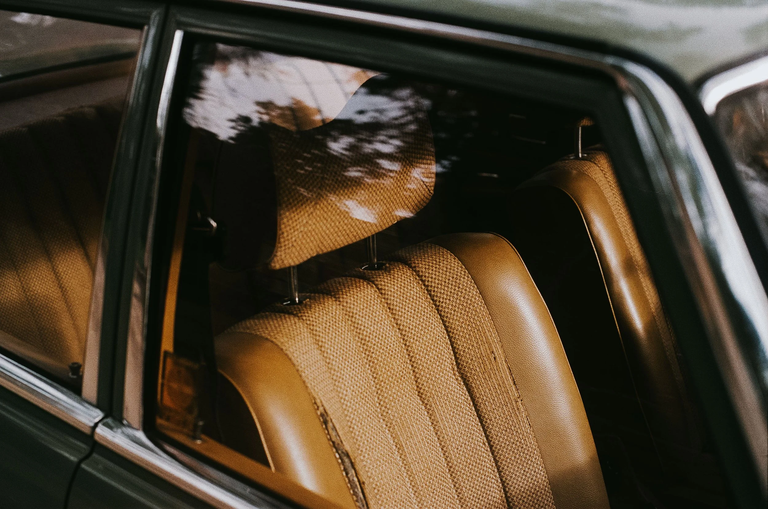 Close-up view of the interior of a vintage car, showing the beige leather seats with a plaid pattern design and the car's window with reflections of trees.