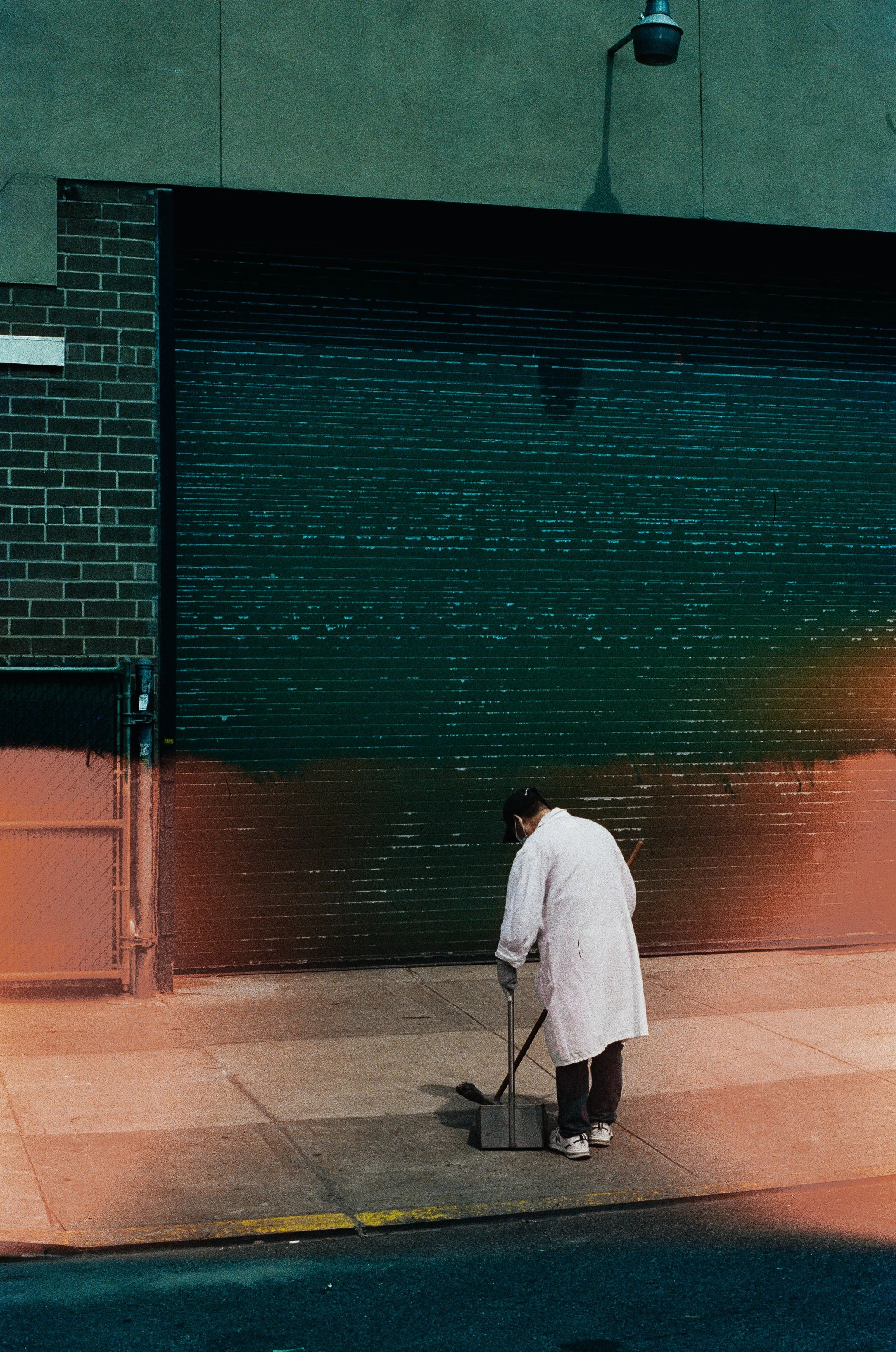Person wearing a white coat and mask sweeping sidewalk outside a building with a closed dark-colored garage door.