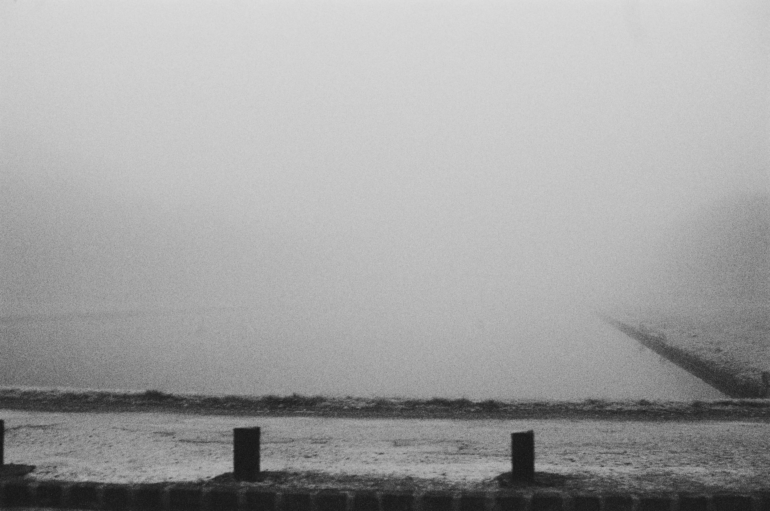 Black and white photo of a snow-covered beach with a pier extending into the water on the right side, and several small posts along the shoreline.