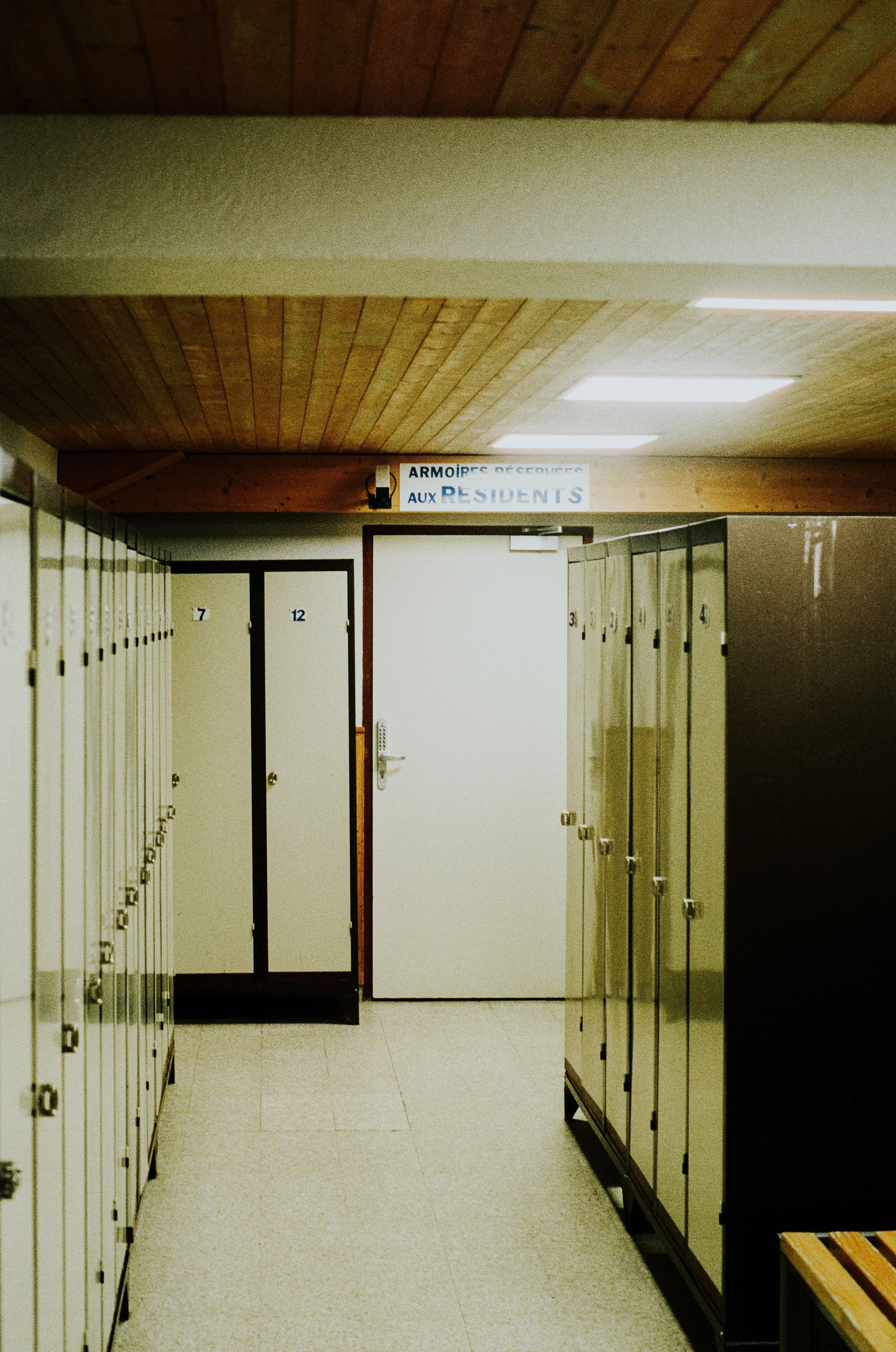 A hallway with beige lockers along both sides, a white door at the end, and a sign above the door that reads 'Armoires réservées aux résidents'.