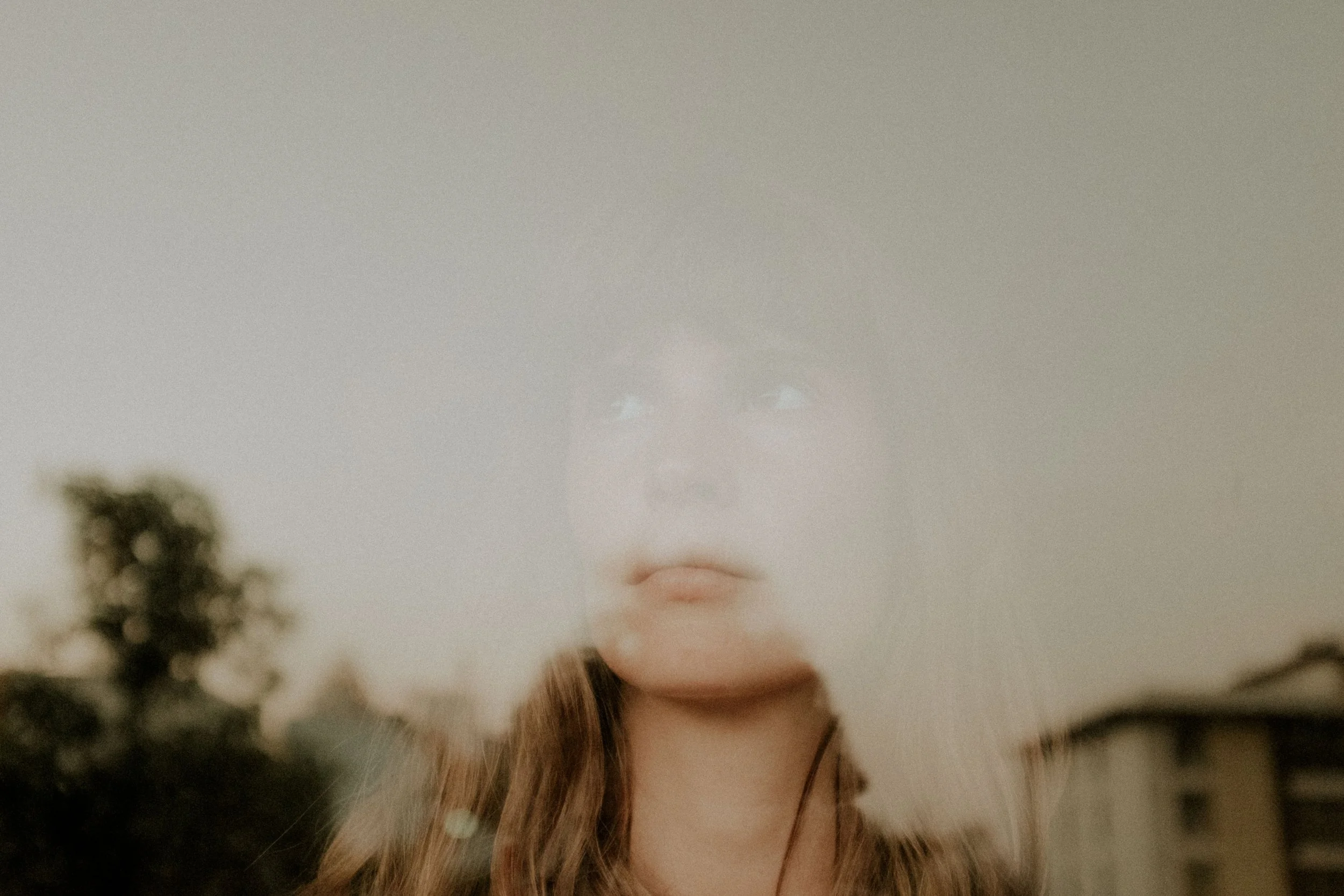 A double exposure photo of a woman's face blended with a cloudy sky and trees, creating a surreal effect.