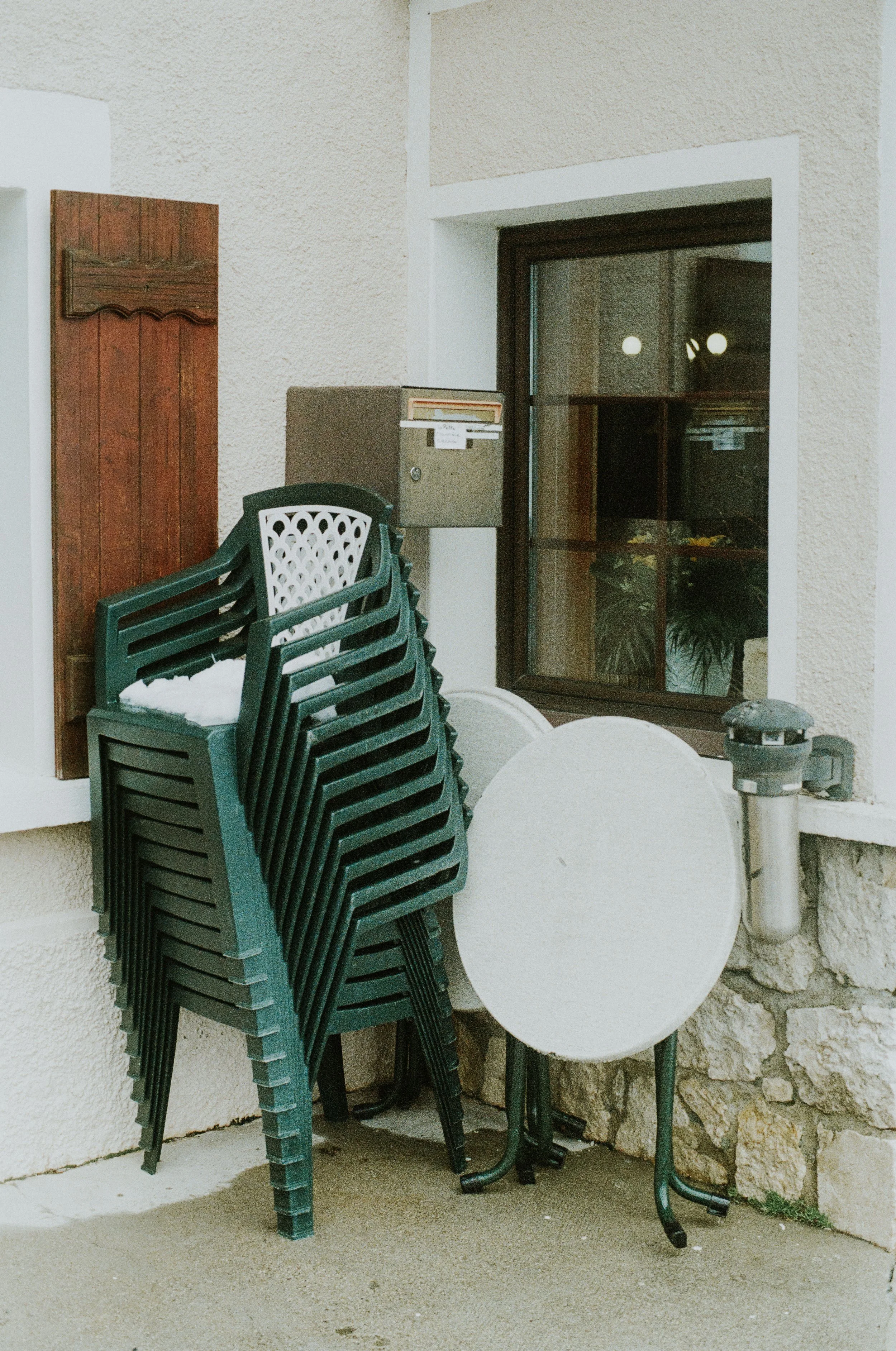 Stack of green plastic chairs, a white round table, a window, a mailbox, a vent pipe, and a wooden board outside a building.