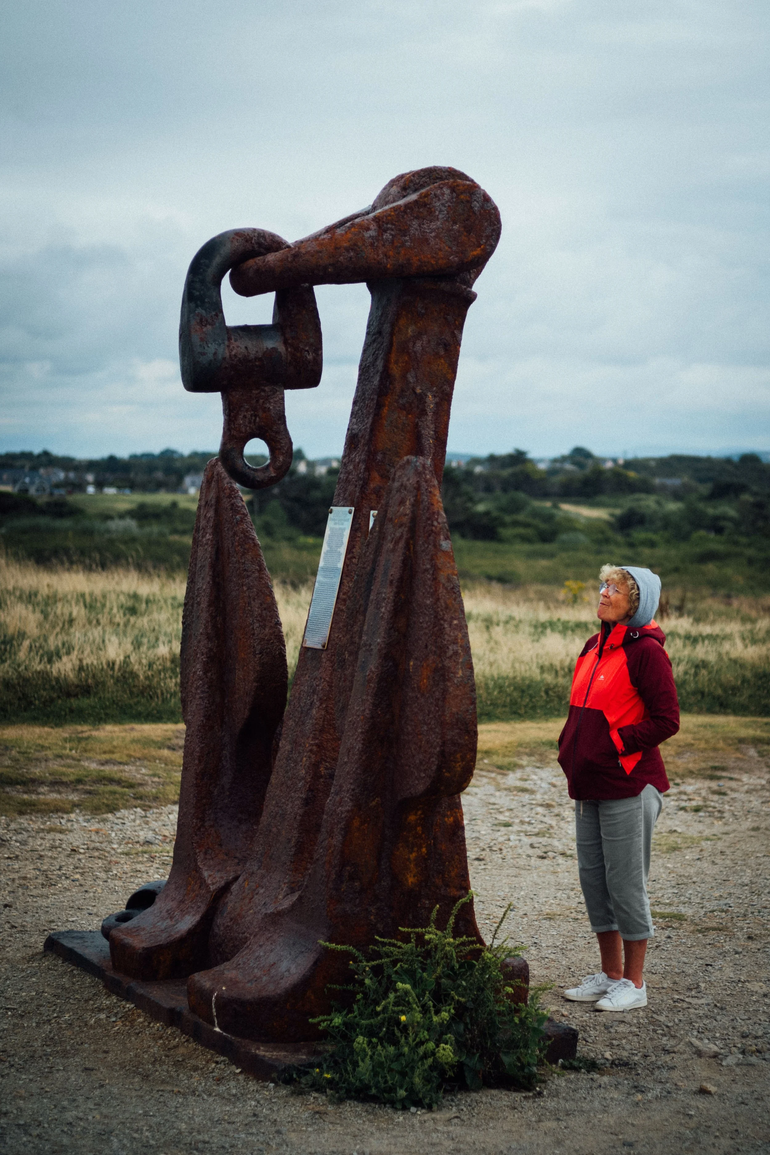 A woman in a red and black jacket, grey pants, and a grey hoodie stands next to a large, rusted metal sculpture of an anchor outdoors in a grassy landscape with hills in the background.