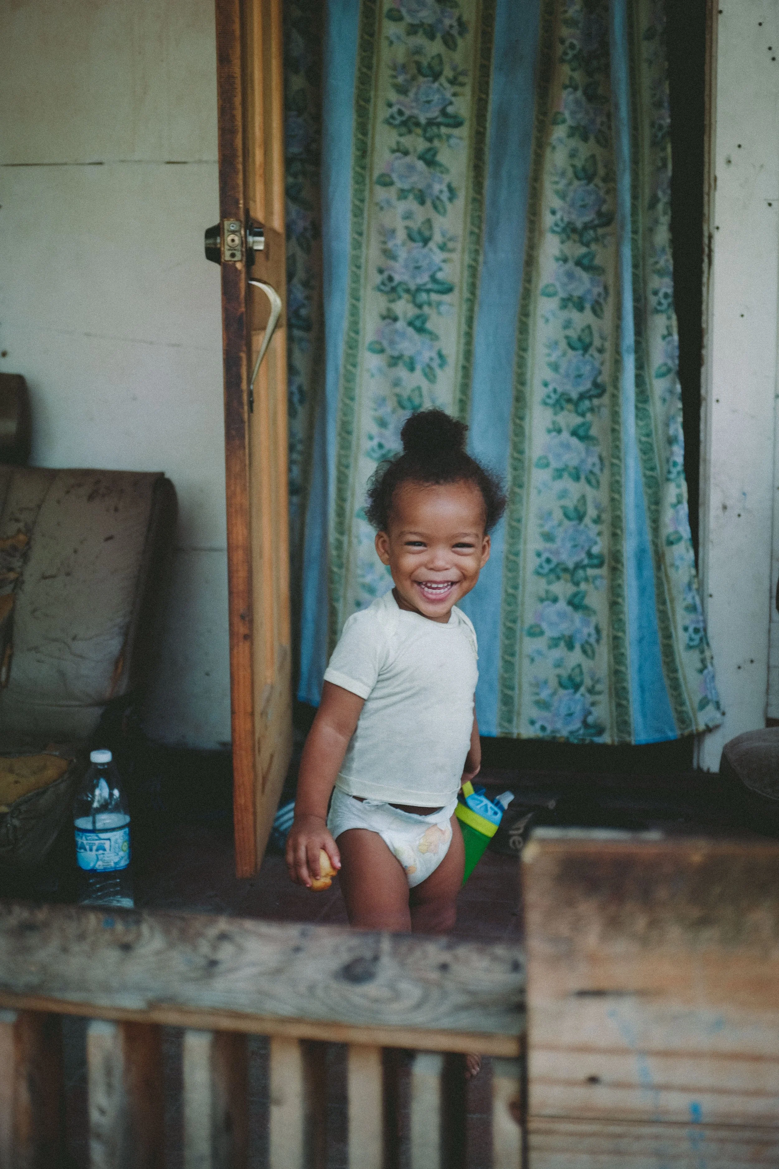 A young girl with curly hair tied up in a bun, smiling widely, standing indoors near a wooden structure and a curtain with a floral pattern.