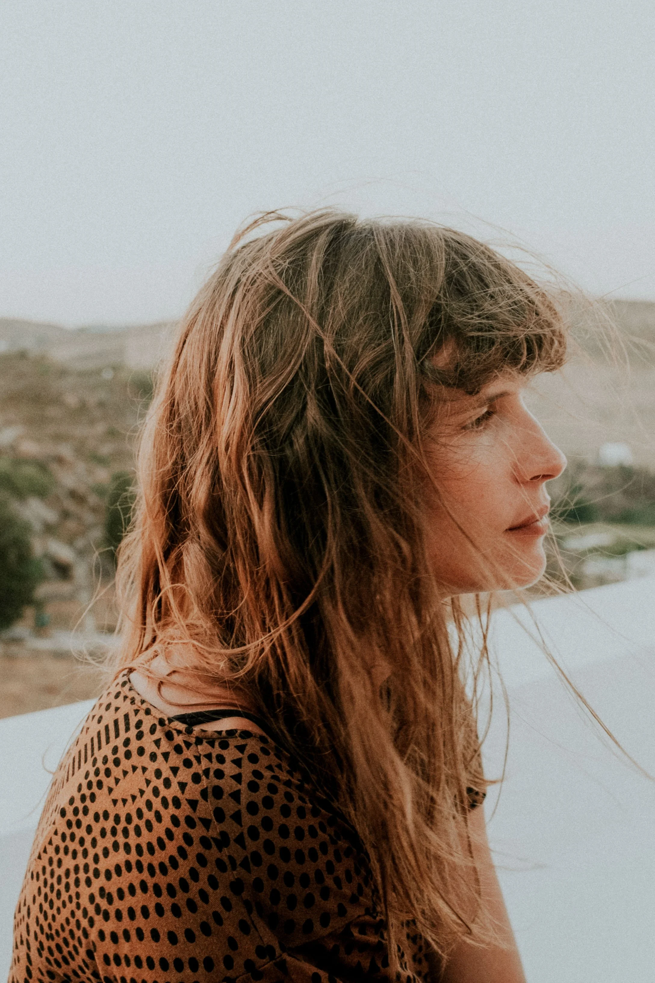 A woman with long, wavy hair gazes to the right outdoors, with a distant landscape of hills and a light sky in the background.
