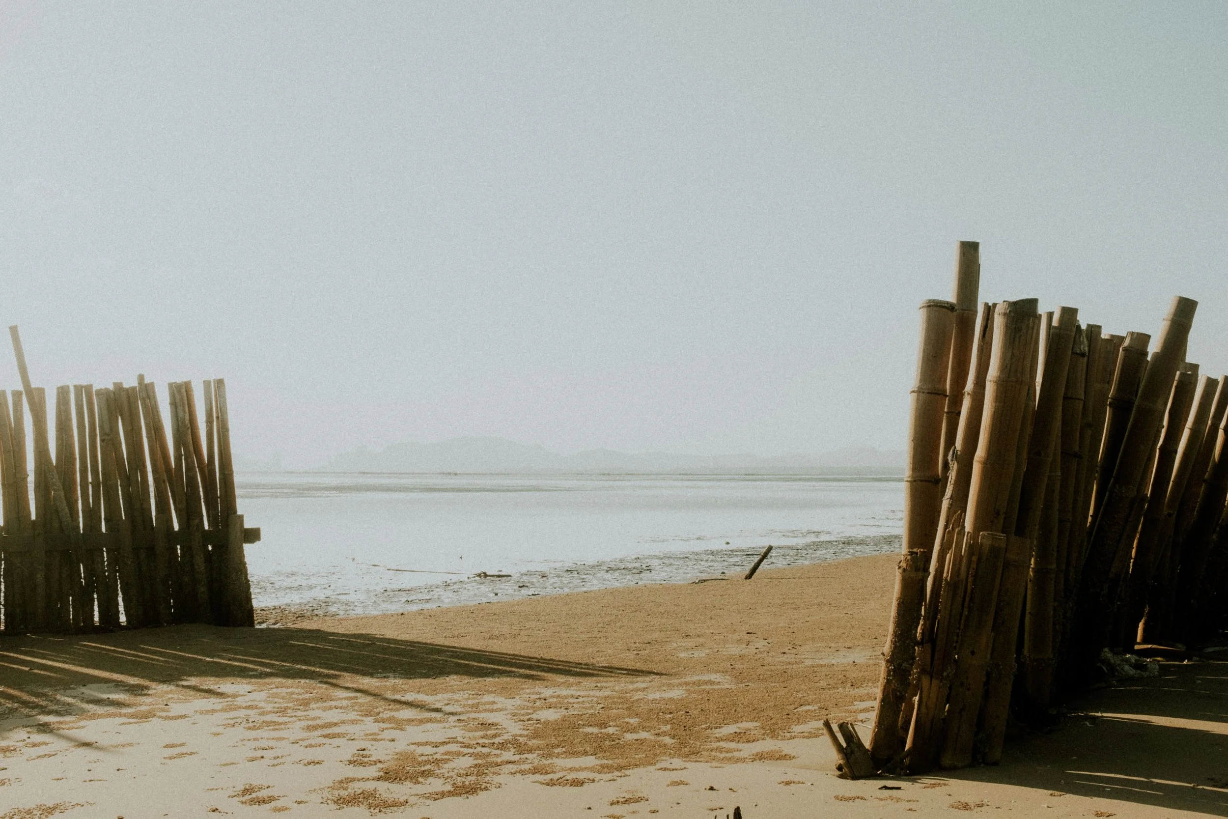 Bamboo fences on a sandy beach near shallow water, with a hazy sky and distant landforms on the horizon.