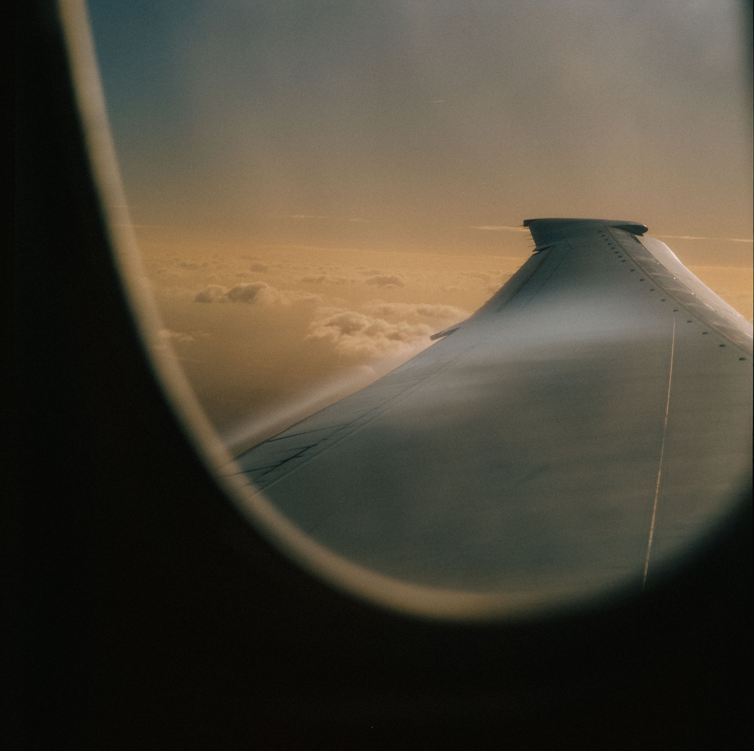 View of an airplane wing through the window during flight, with clouds below and a golden sky at sunrise or sunset.