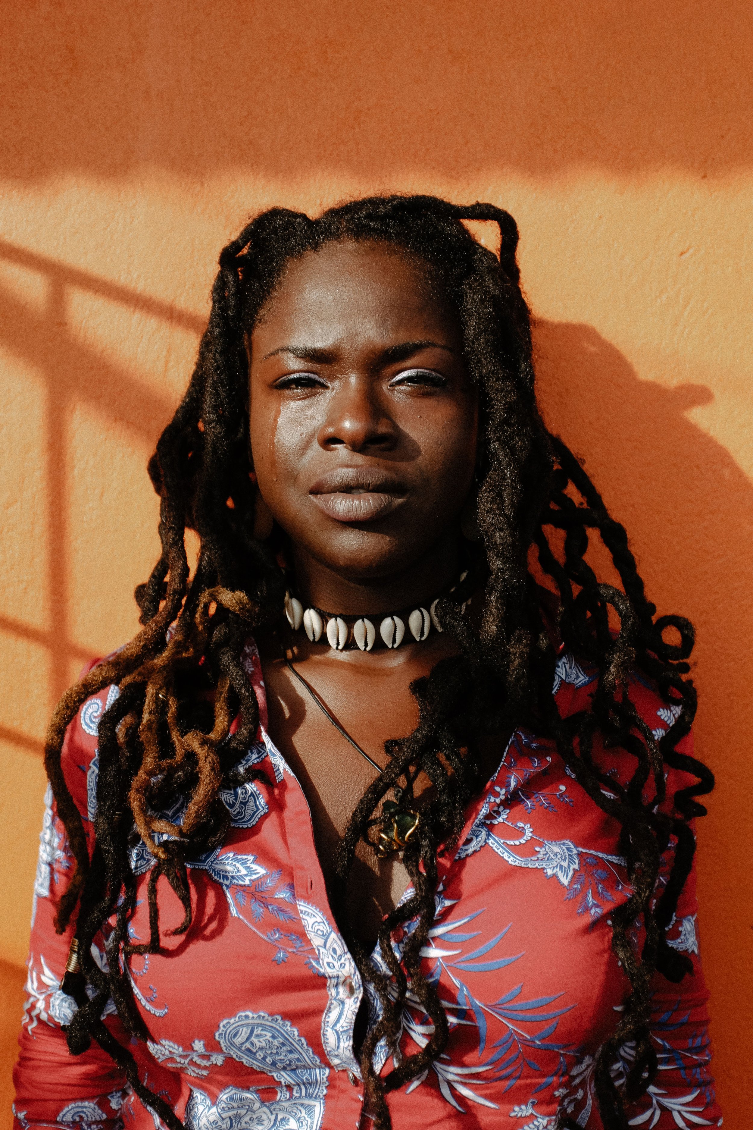 A woman with dark braided hair styled in dreadlocks, wearing a shell necklace, a necklace with a pendant, and a colorful patterned blouse, standing against an orange wall with shadows.