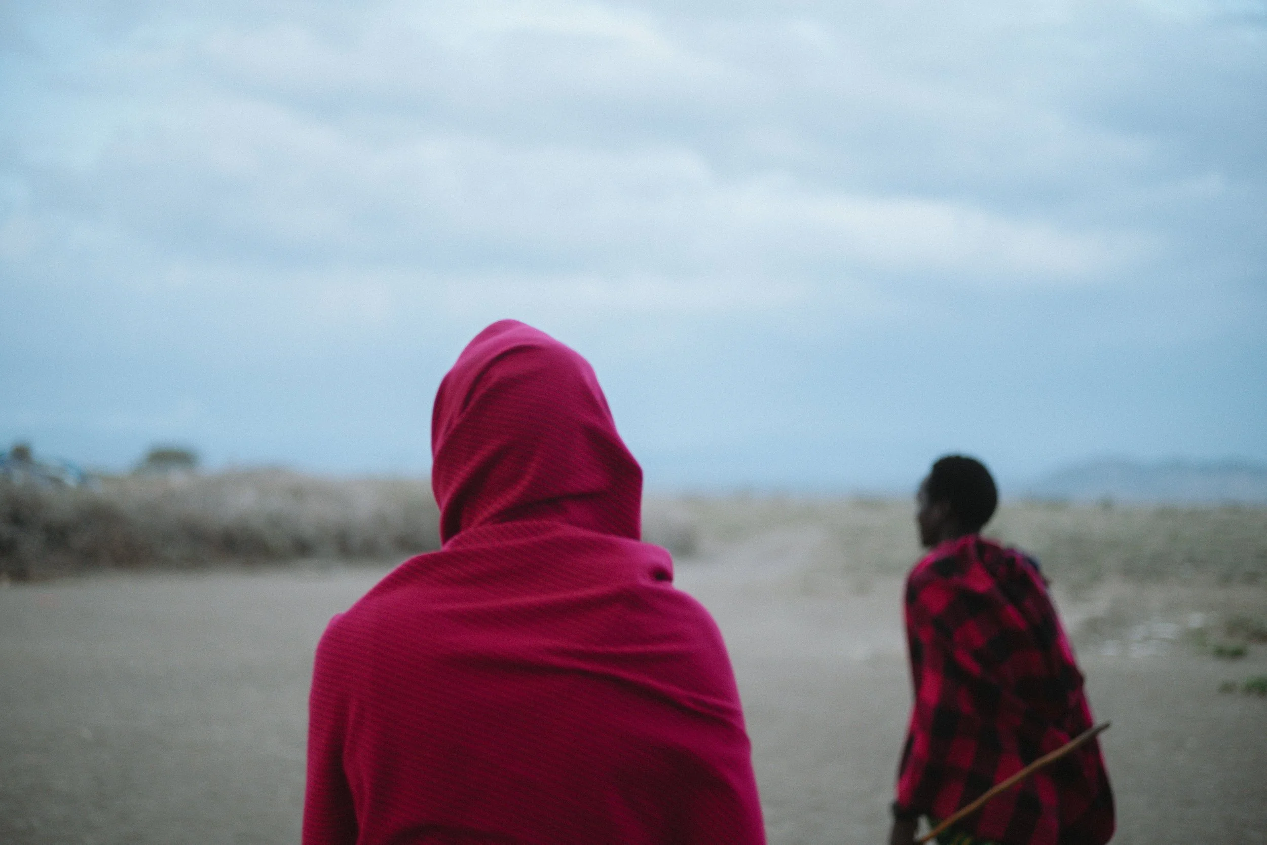 Two people sitting on a sandy beach, one wearing a pink hooded garment, the other wearing a red and black plaid shirt, with overcast sky overhead.